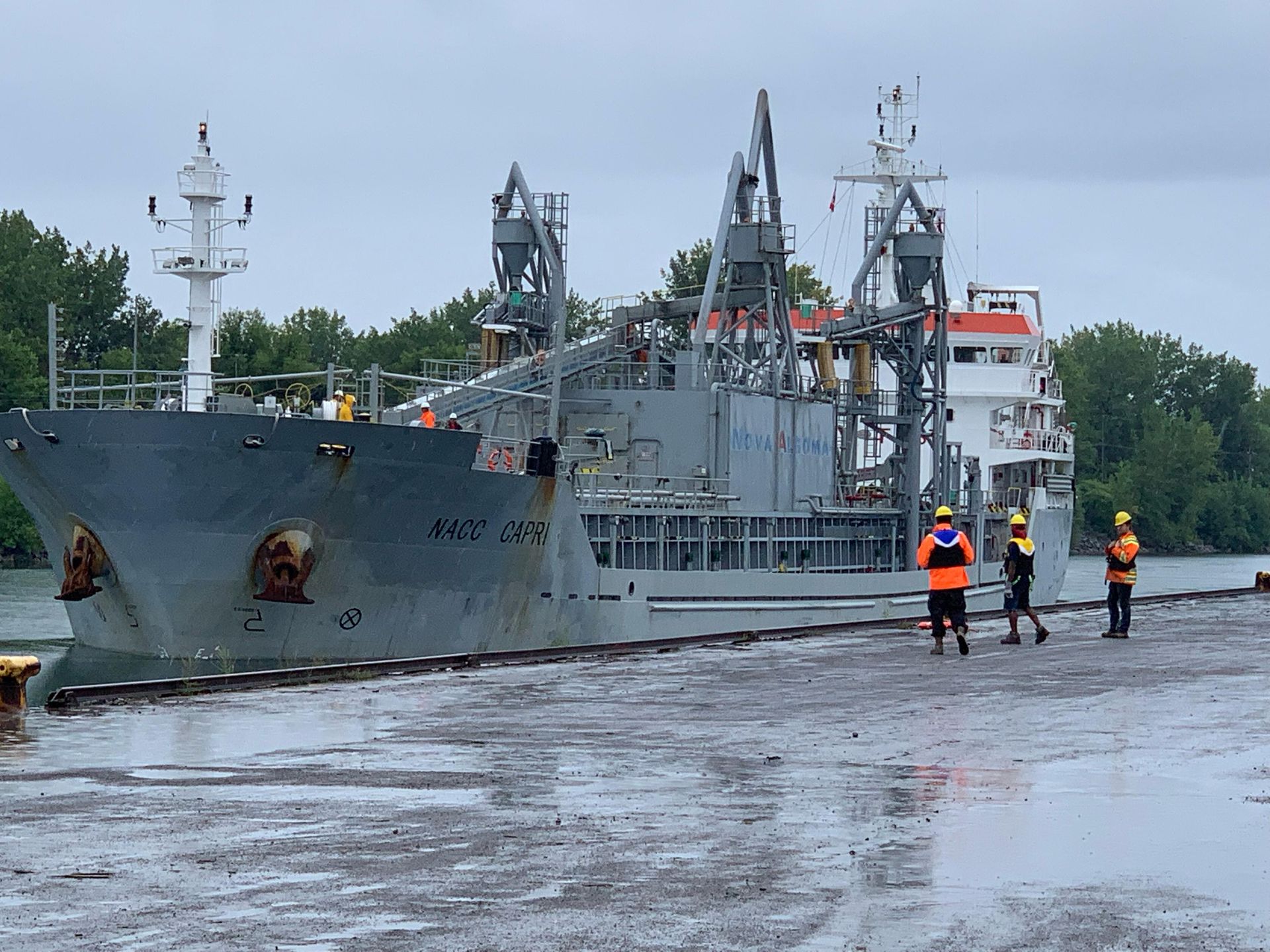 Crane lifting a white container near a blue ship. A worker in safety vest watches. Outdoors.