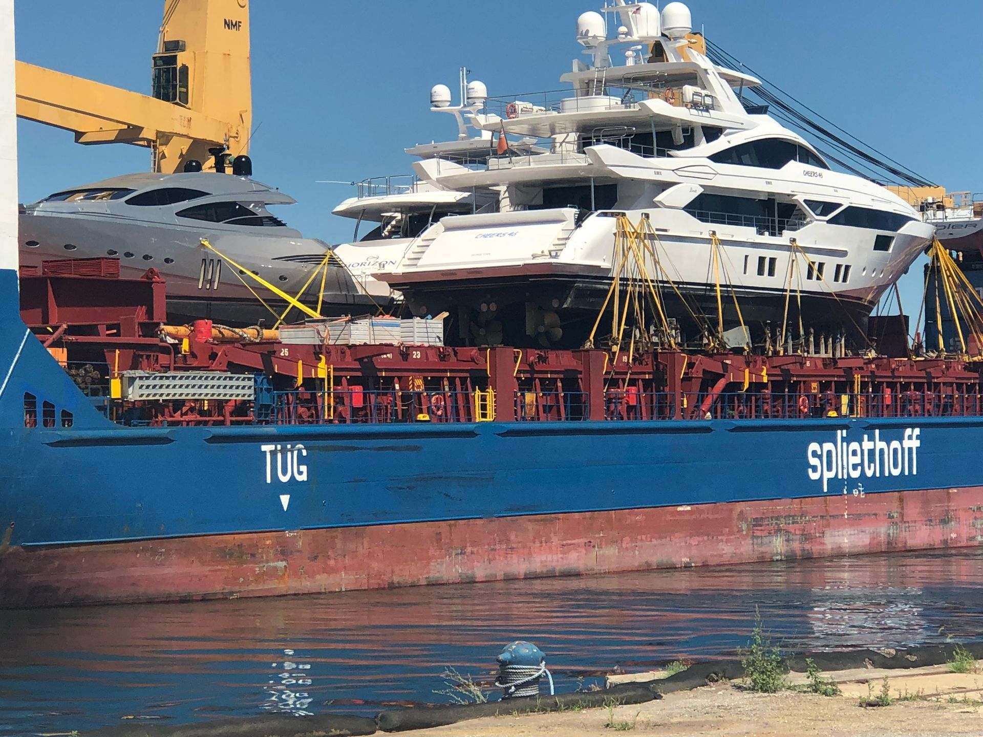 Two large yachts on a blue cargo ship. Cranes and support cables secure the yachts. Water and sky in background.