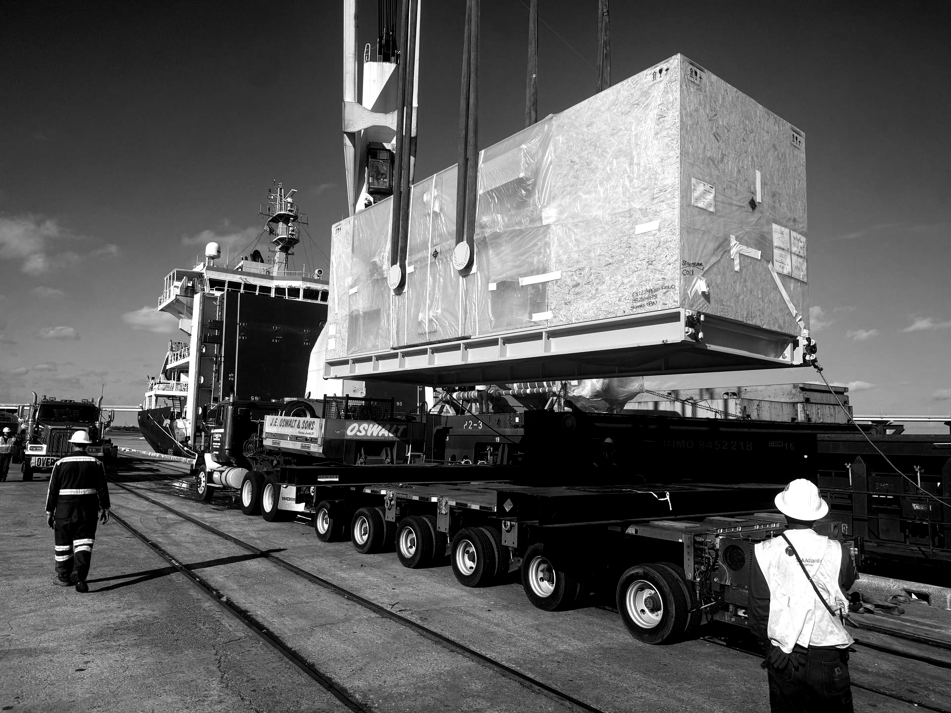 Oversized cargo loaded on a long trailer, with workers guiding it on a road under a cloudy sky