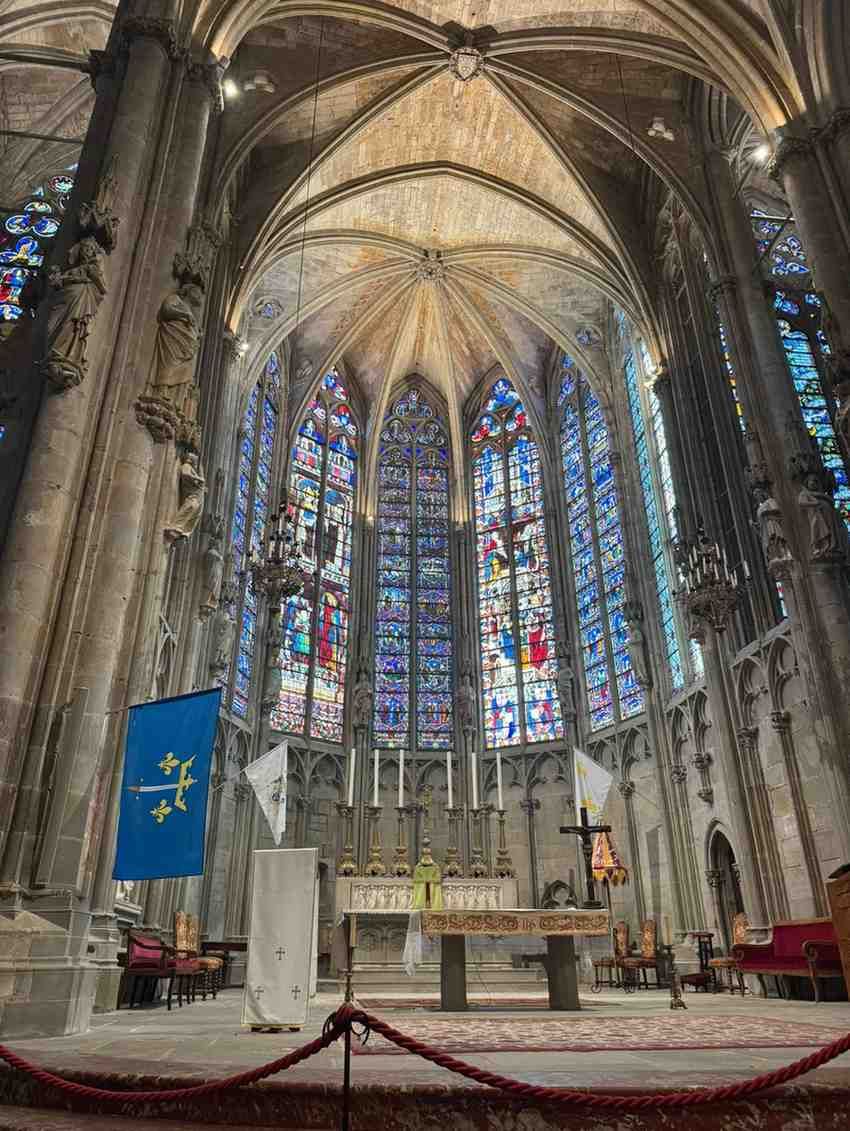 Interior de una catedral gótica con vidrieras, techo abovedado, altar y estandarte azul colgando a la izquierda.