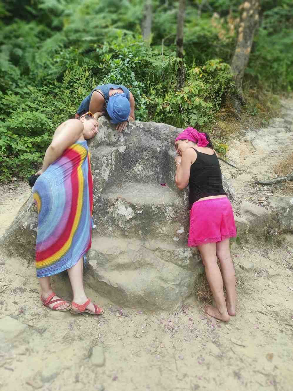 Dos personas apoyadas en una gran roca en un sendero arenoso; una lleva una toalla de colores del arcoíris.