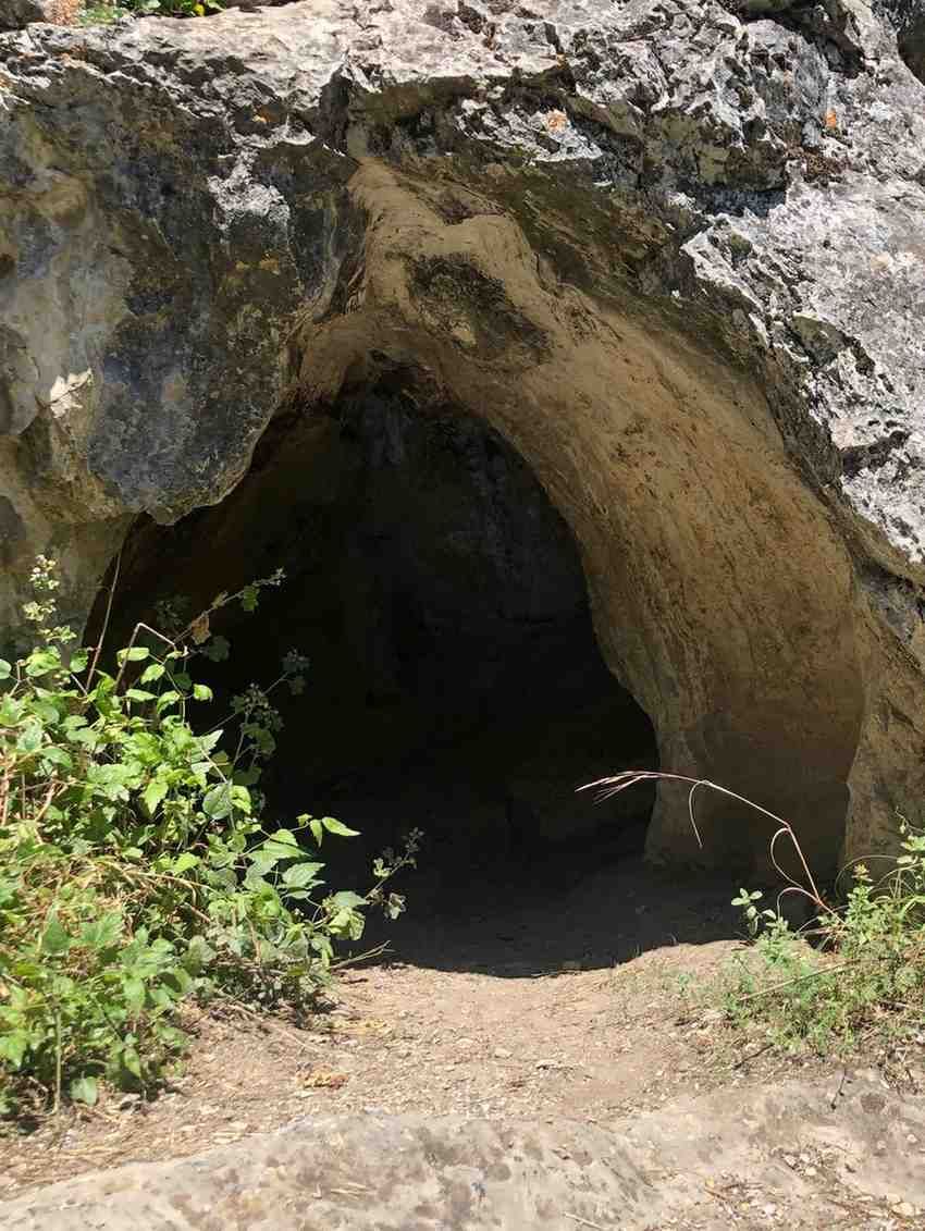 Entrada rocosa a una cueva con una abertura oscura y suelo arenoso rodeado de escasa vegetación.