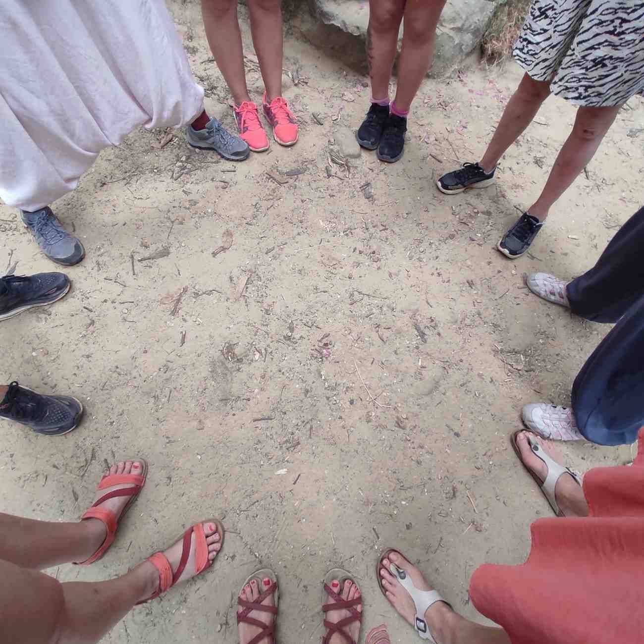 Personas de pie formando un círculo sobre un terreno arenoso, mostrando zapatos y sandalias.
