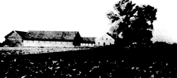 Ländliche Landschaft in Schwarz-Weiß mit einer langen Scheune, Bäumen und einem dunklen Feld im Vordergrund