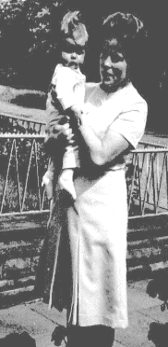 Frau hält ein Kind auf einer Brücke, im Hintergrund Bäume.