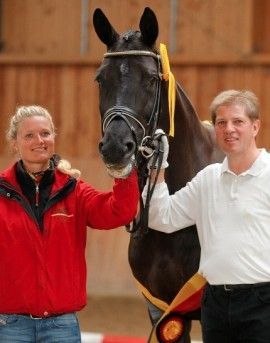 Eine Frau in roter Jacke und ein Mann in weißem Hemd stehen neben einem schwarzen Pferd mit einer Schleife in einem Stall.