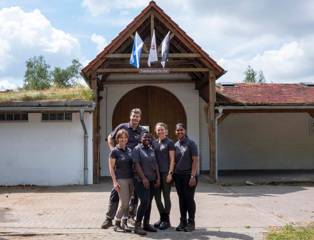 Fünf Personen in dunklen Hemden posieren vor einem weißen Gebäude mit einem Holzbogen und darüber hängenden Fahnen.