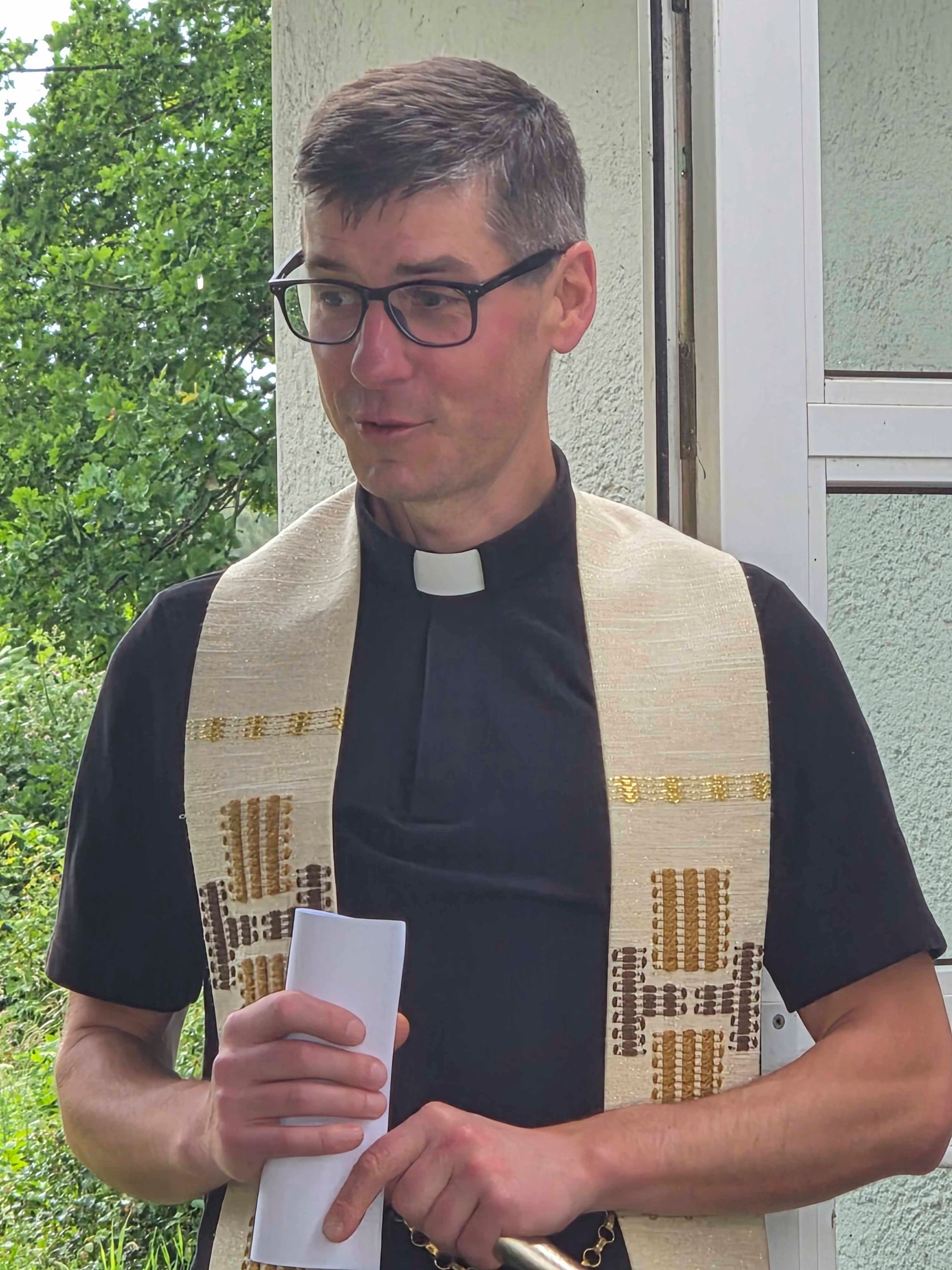 Ein Geistlicher in schwarzem Hemd und weißer Stola hält drinnen in der Nähe eines Fensters Papiere in der Hand.
