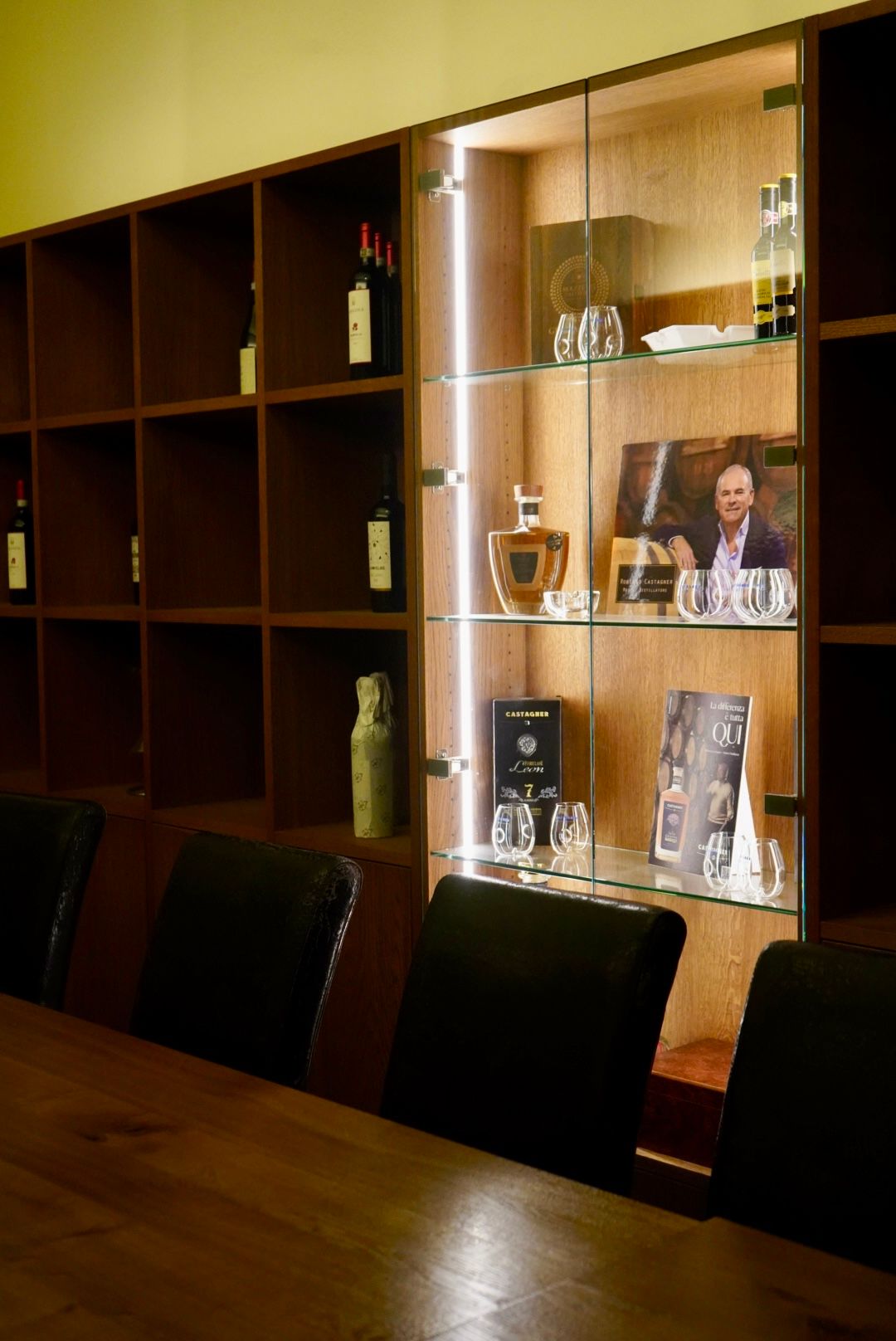 Holzwand mit Regalen, Glasvitrine mit Spirituosenflaschen und Gläsern. Tisch mit schwarzen Stühlen davor.