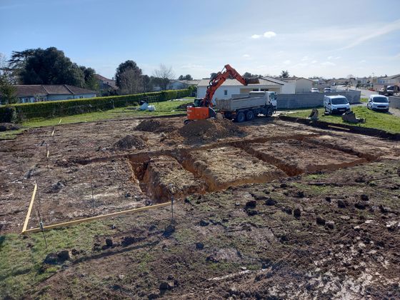 Chantier de construction avec une excavatrice creusant une grande tranchée rectangulaire dans un terrain en terre.