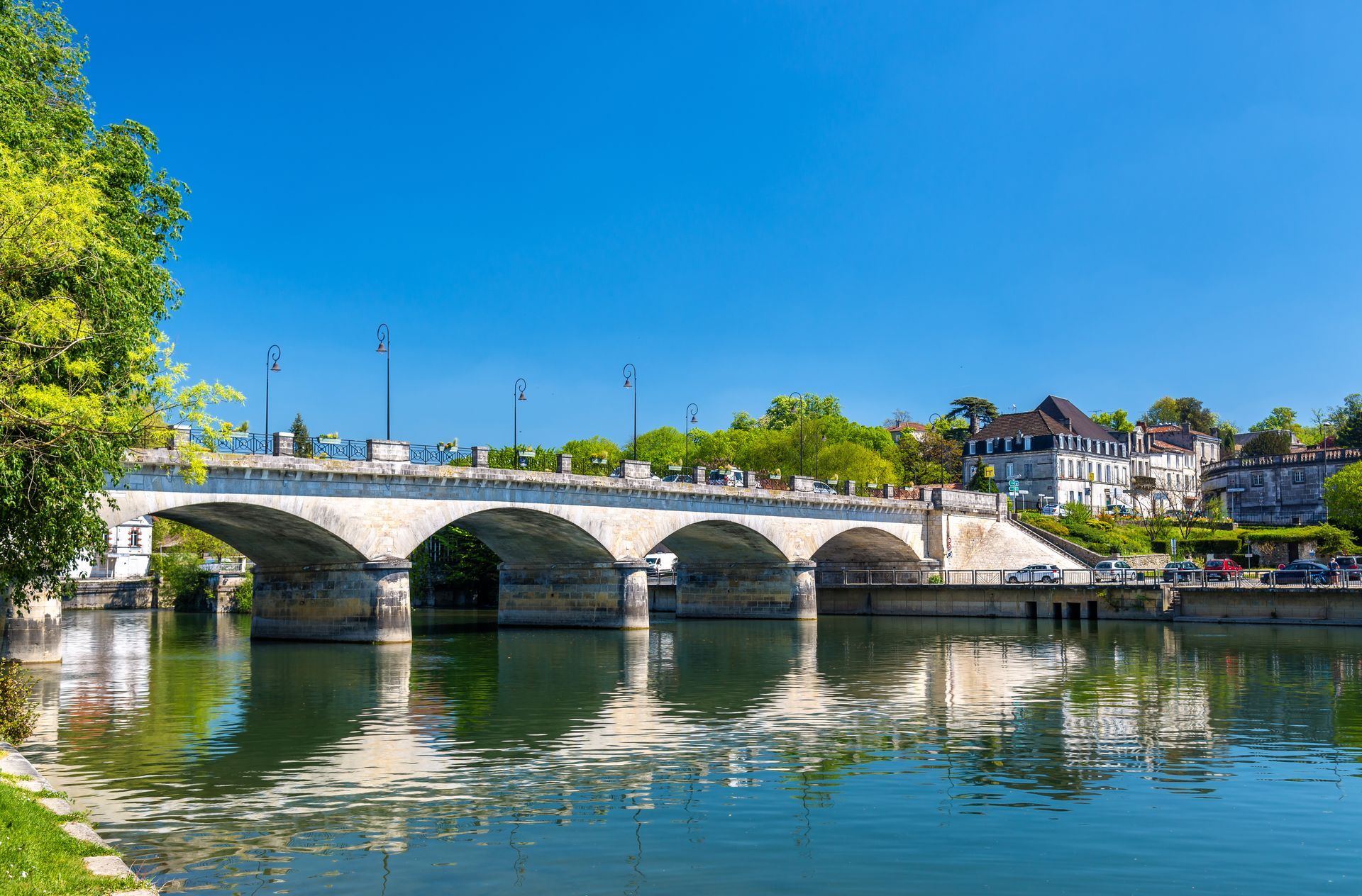 Pont de pierre enjambant une rivière calme, sous un ciel bleu, avec des arbres et des bâtiments le long du rivage.