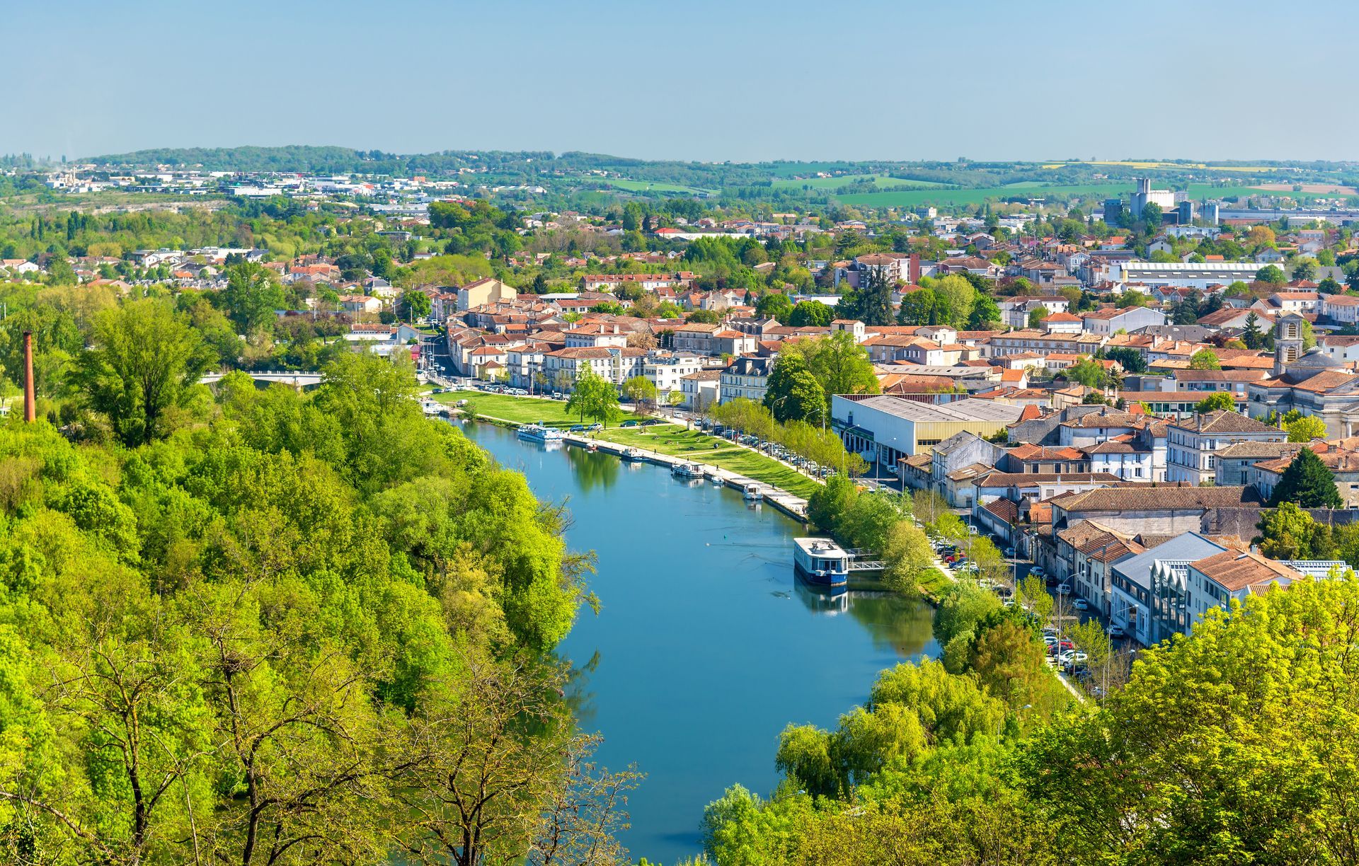Vue aérienne d'une rivière serpentant à travers une ville verdoyante, avec des maisons et des bateaux sous un ciel dégagé.