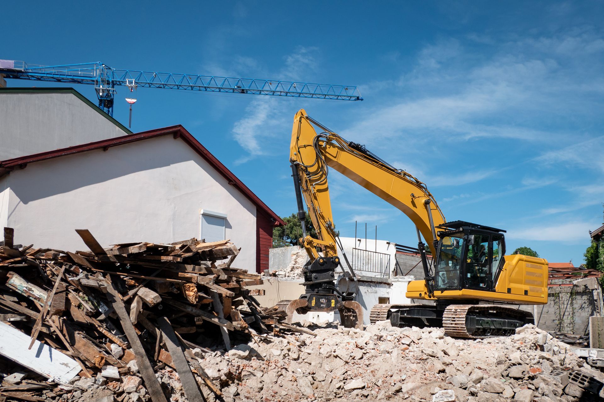 Une pelleteuse jaune démolit des gravats à côté d'une maison blanche sous un ciel bleu.
