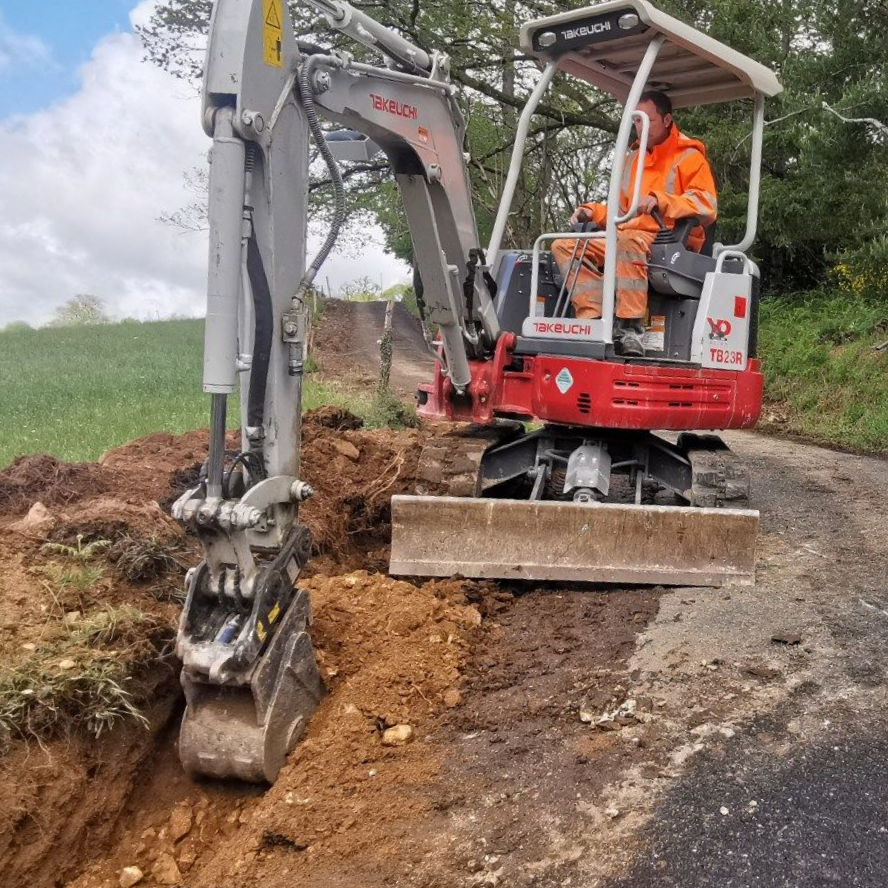 Une excavatrice creuse un fossé en bordure de route de gravier, avec un ouvrier en tenue de sécurité orange dans la cabine.