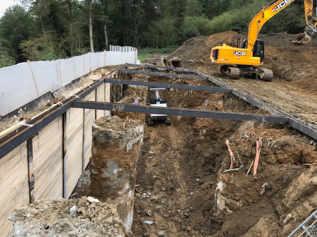 Chantier de construction avec une excavatrice à côté d'une tranchée profonde, des supports en acier et des travaux de mur de soutènement