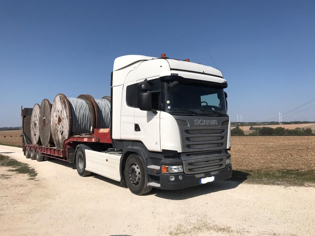 Un semi-remorque blanc transporte de grandes bobines de câble sur une remorque à plateau dans un paysage rural sous un ciel dégagé.