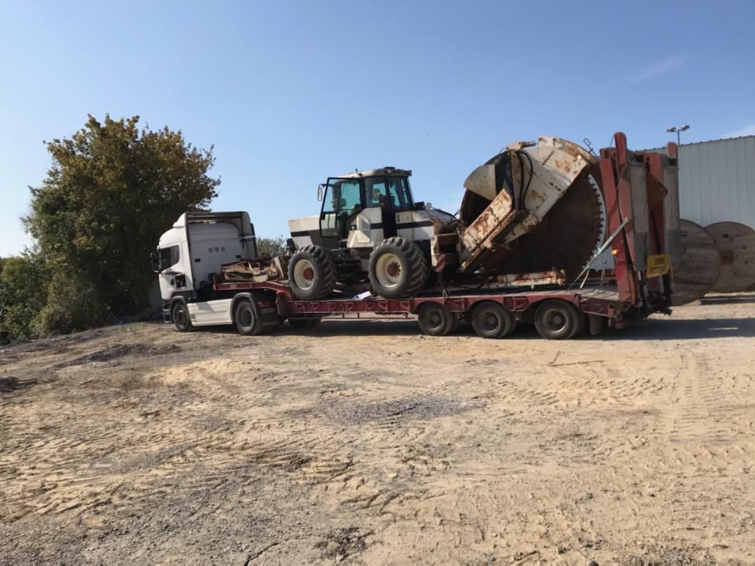 Un tracteur sur une remorque à plateau sur un chantier poussiéreux, avec une grande bétonnière à proximité.