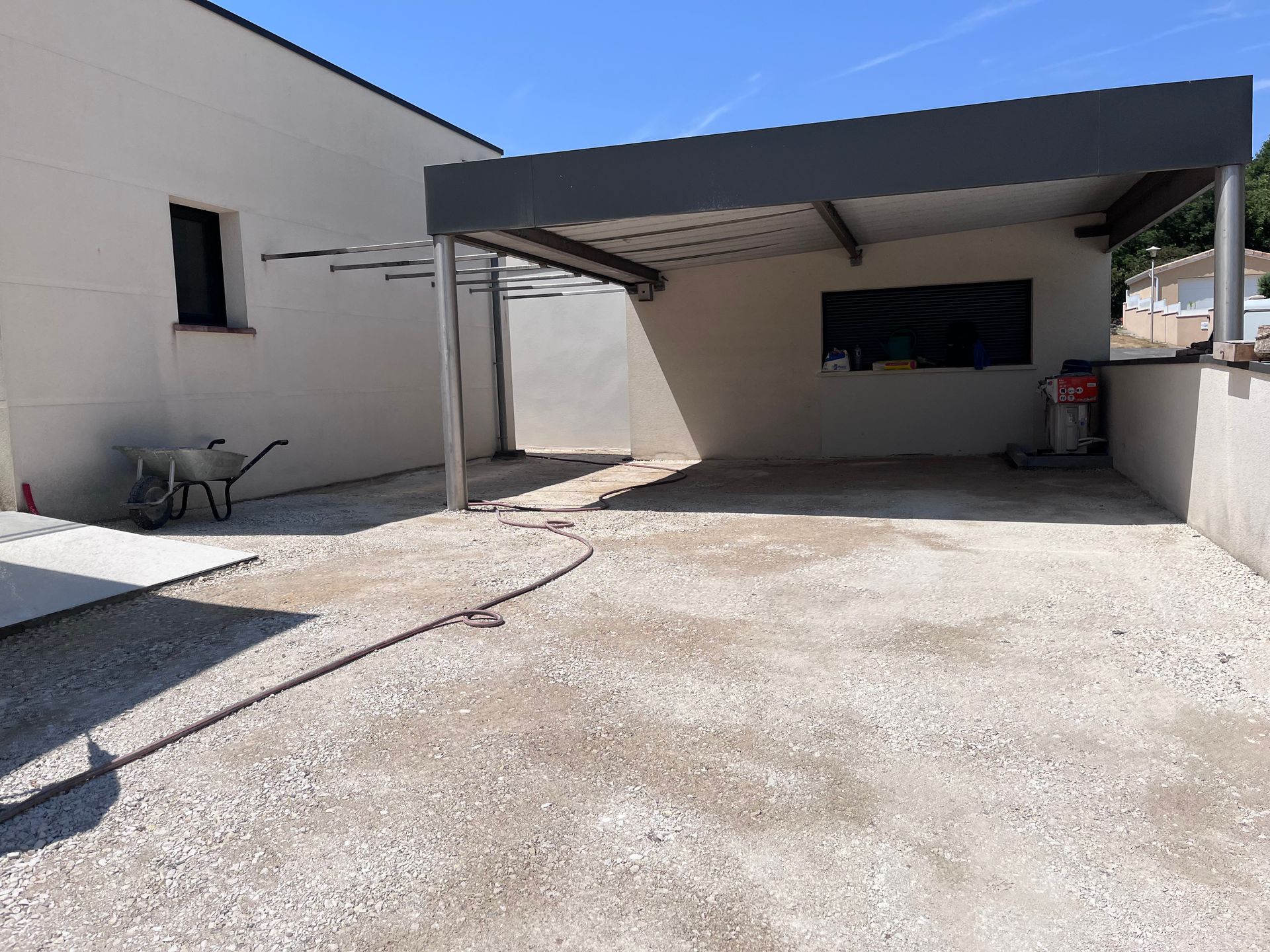 Cour en béton aux murs blancs, un abri de voiture en métal et une brouette sur la gauche.