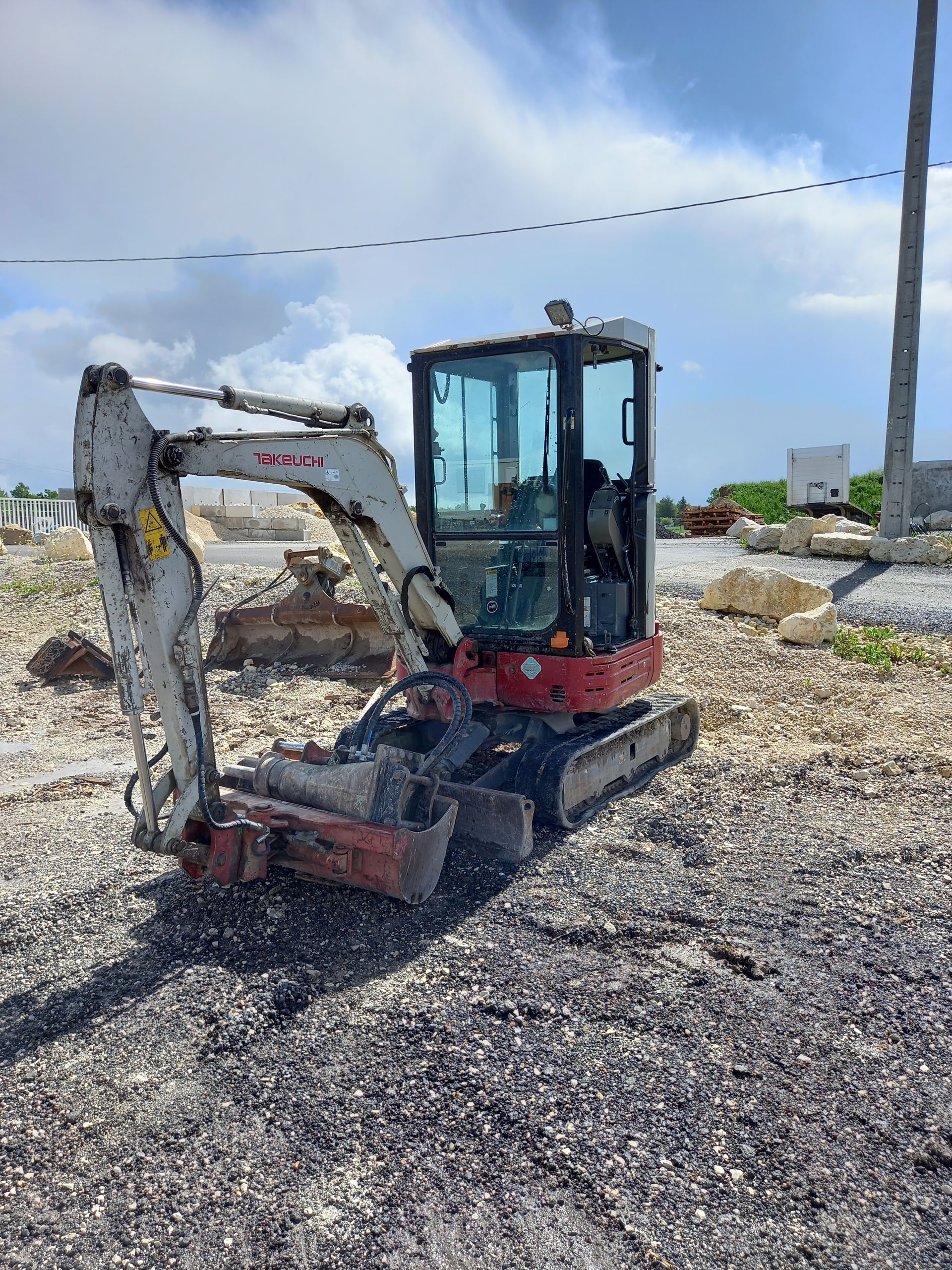 Minipelle blanche sur un terrain gravillonné sous un ciel nuageux, bras levé à côté d'un chantier.