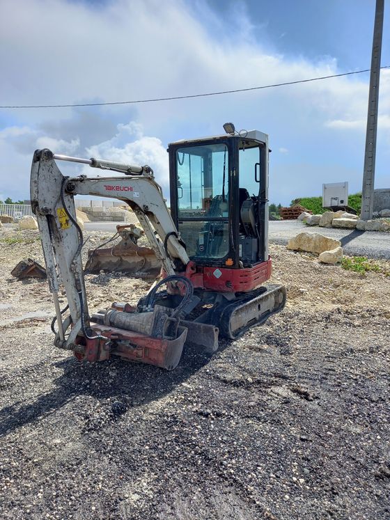 Minipelle blanche sur un terrain gravillonné sous un ciel nuageux, bras levé à côté d'un chantier.