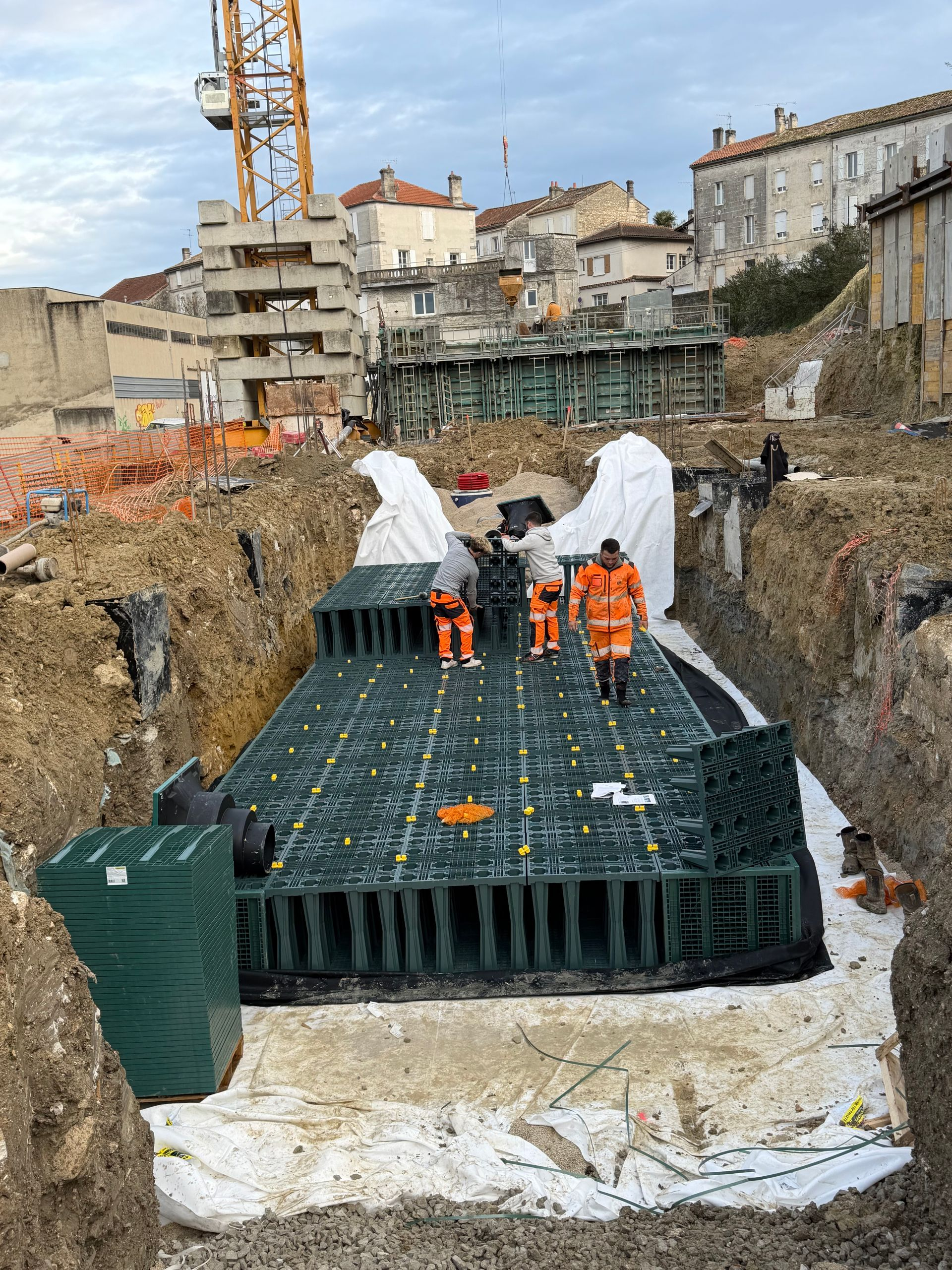 Des ouvriers vêtus de gilets orange installent des bacs de drainage noirs sur un chantier de terrassement.