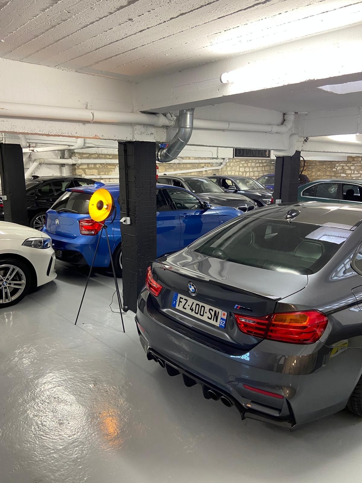 Voitures garées dans un garage, principalement des BMW. Voitures grises et bleues bien visibles. Feu jaune et sol gris.