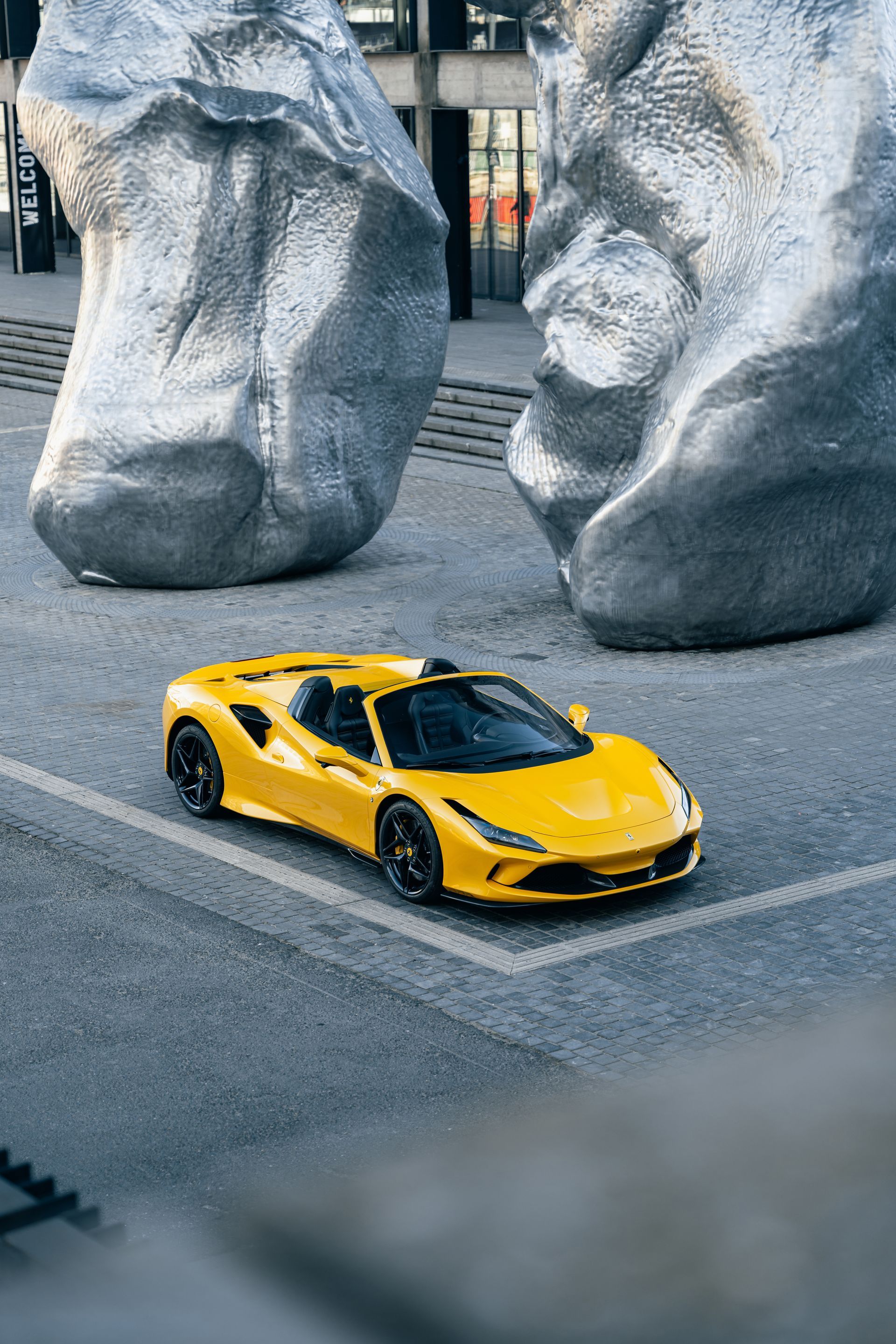 Voiture de sport Ferrari jaune garée sur des pavés, entre deux grandes sculptures grises.