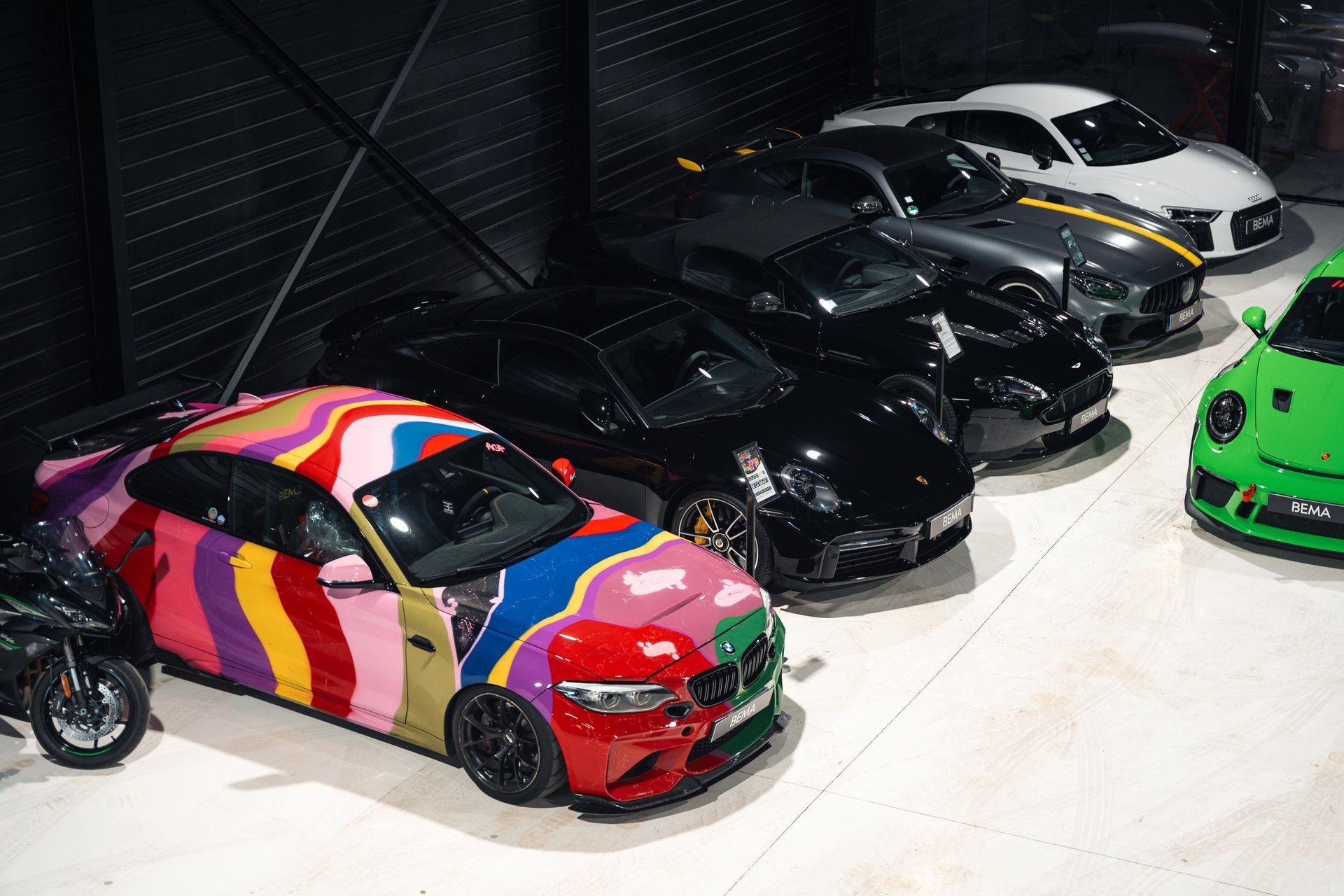 Voitures garées dans un garage ; une BMW aux couleurs vives, plusieurs voitures noires et une Porsche verte sont visibles.