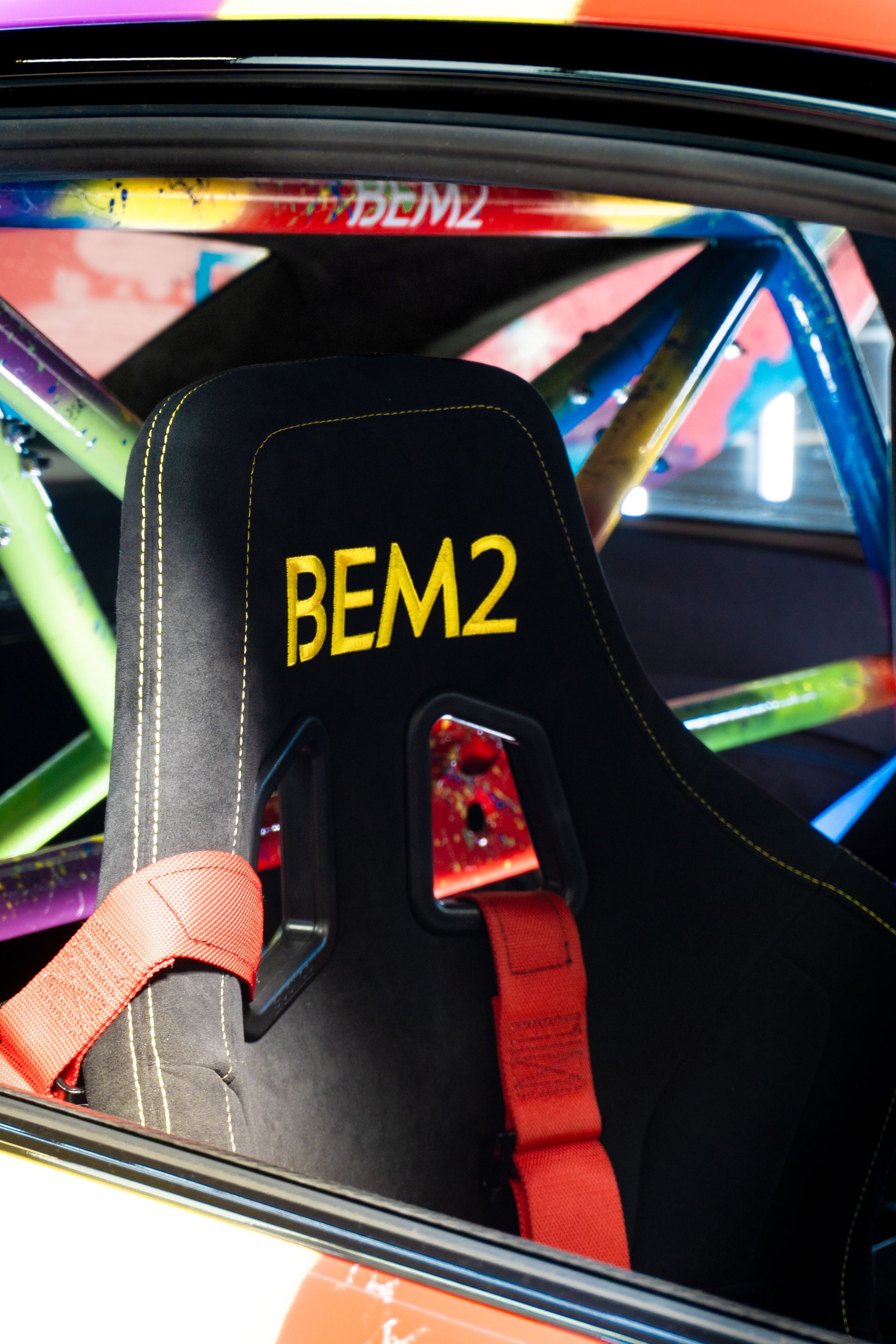 Siège de course noir avec lettrage jaune « BEM2 » et sangles de harnais rouges, à l'intérieur d'une voiture avec un arceau de sécurité coloré.