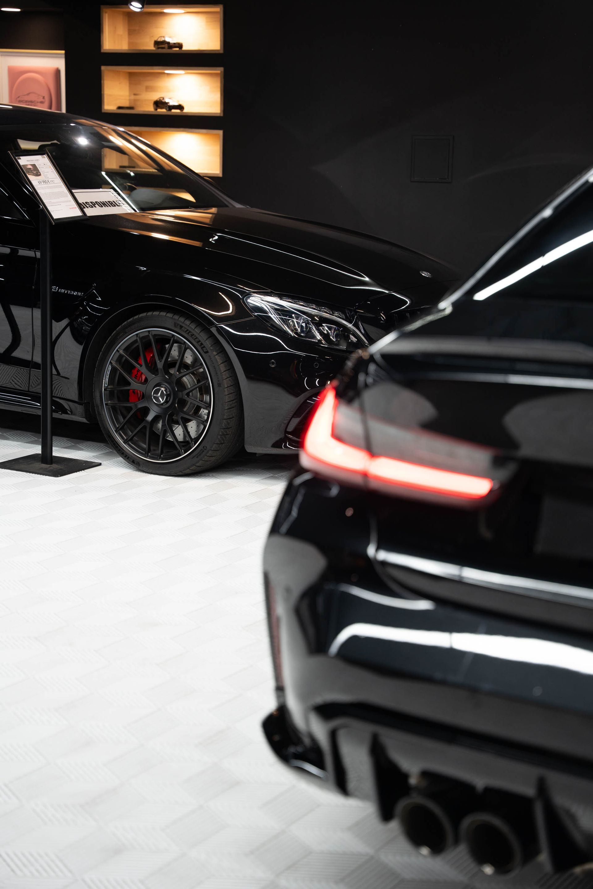 Deux voitures noires exposées dans un showroom. Les feux arrière et le pot d'échappement de l'une sont visibles ; l'autre est partiellement visible.