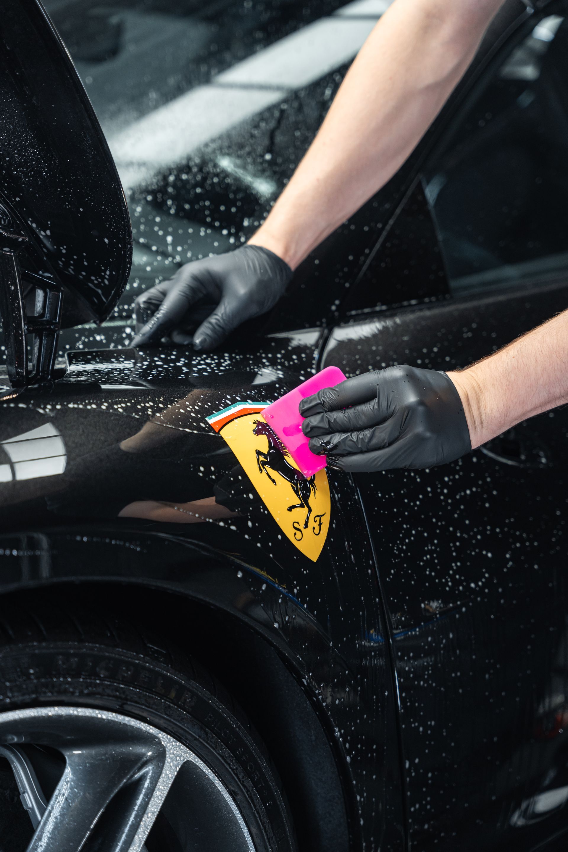 Des mains portant des gants noirs lissant un outil rose sur un emblème Ferrari noir mouillé.