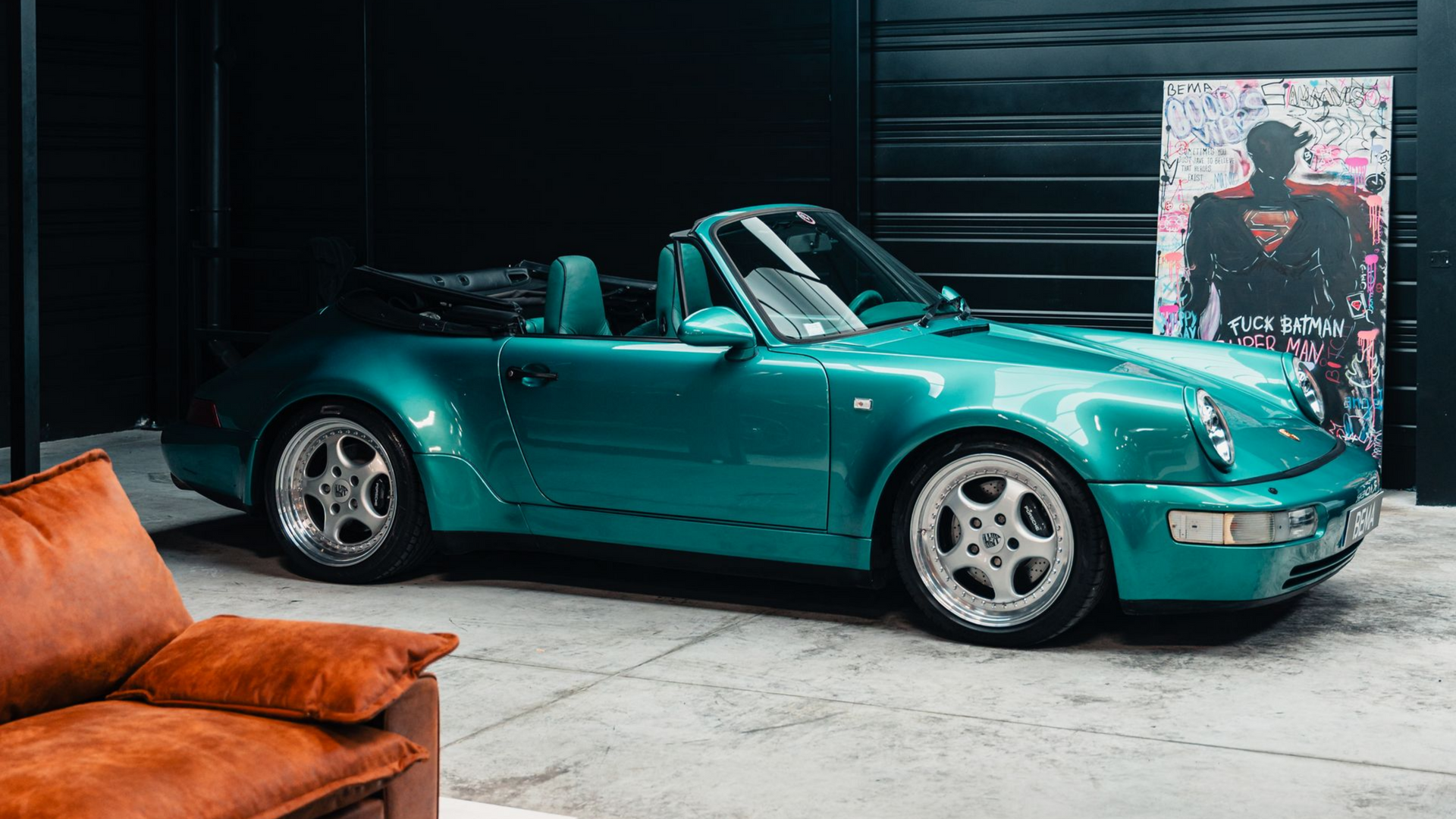 Porsche 911 décapotable turquoise dans un garage sombre avec un canapé, des œuvres d'art et des voitures miniatures.