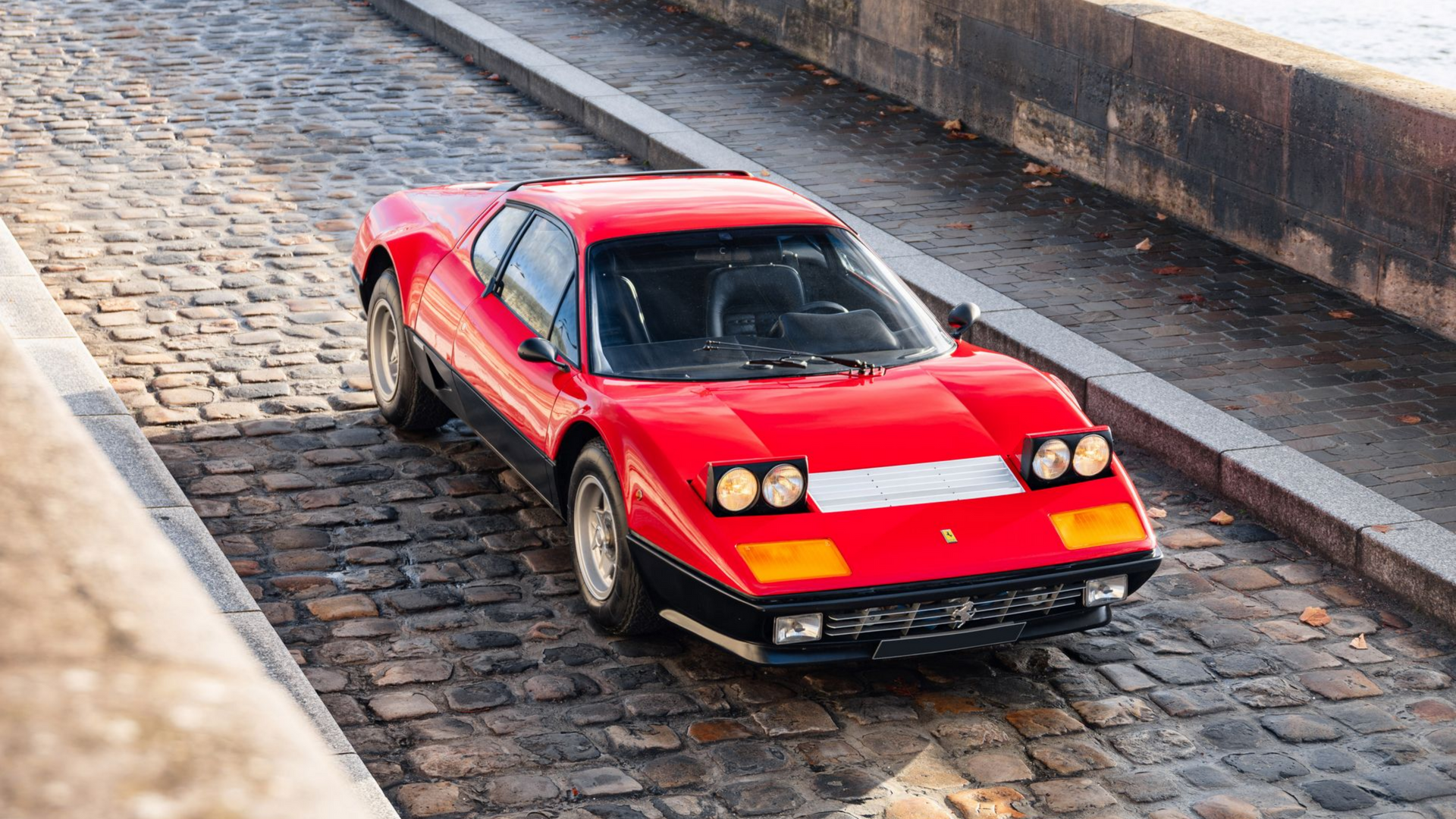 Une Ferrari rouge roule sur une route pavée près d'un pont, par une journée ensoleillée.