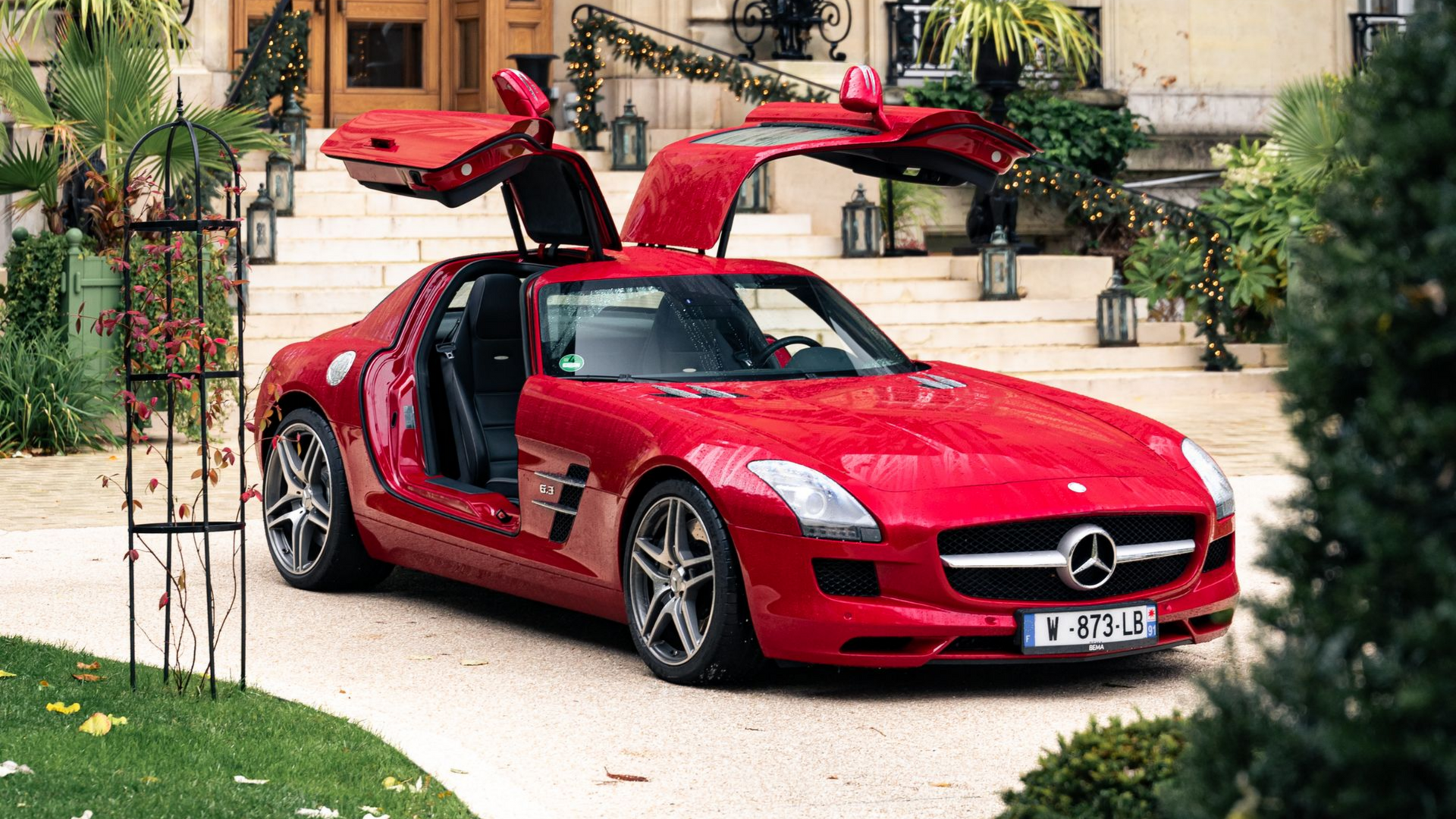 Une Mercedes-Benz rouge à portes papillon est garée devant un grand bâtiment aux portes ouvertes.