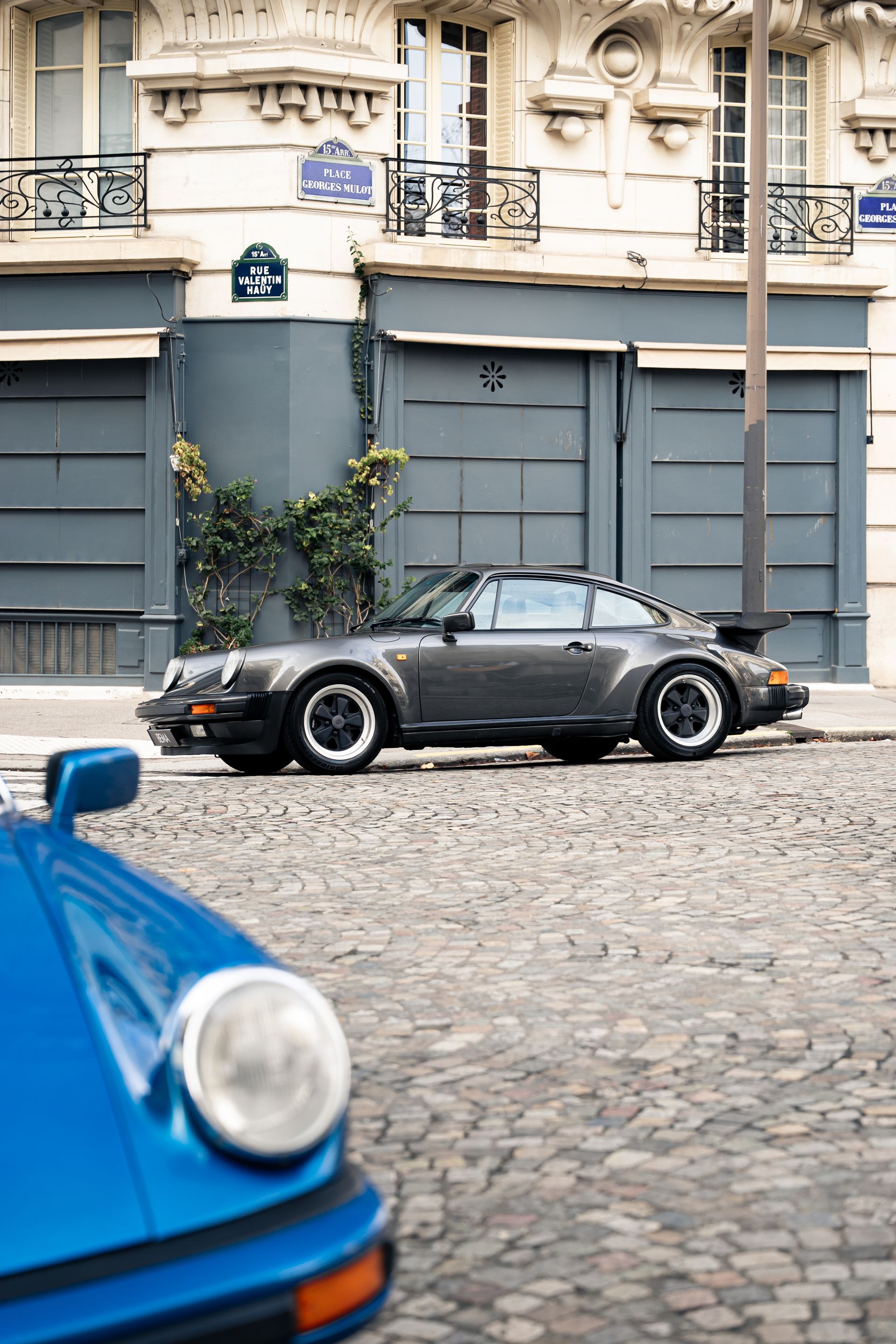 Deux Porsche classiques sur une rue pavée. Une Porsche 911 Turbo grise est garée, et une bleue se trouve au premier plan.