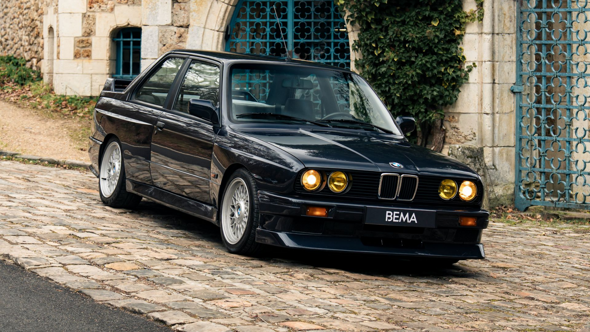 Une BMW E30 M3 noire est garée sur un chemin pavé devant un bâtiment en pierre entouré de végétation.