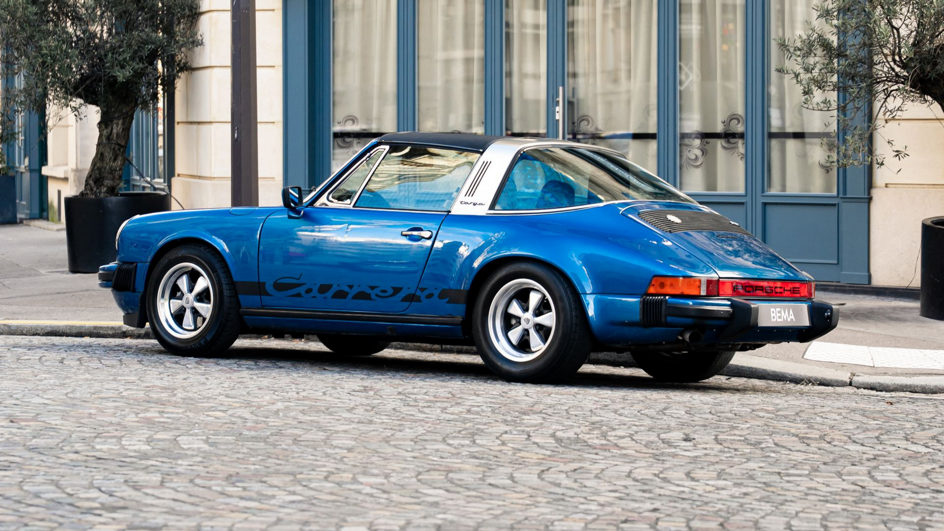 Une Porsche Targa bleue est garée dans une rue pavée devant un immeuble à auvent bleu à Paris.