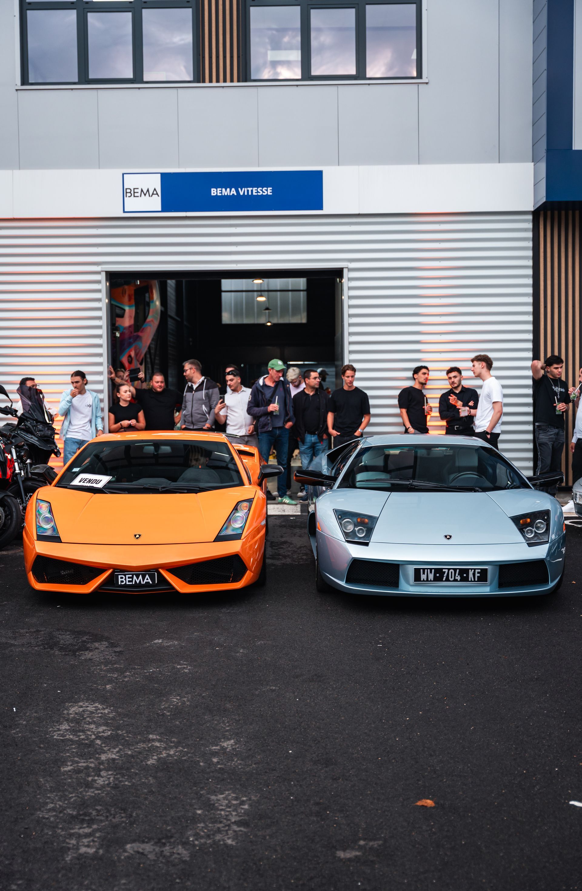 Des Lamborghini orange et argent garées à l'extérieur d'un bâtiment, avec une foule de personnes rassemblées à proximité.