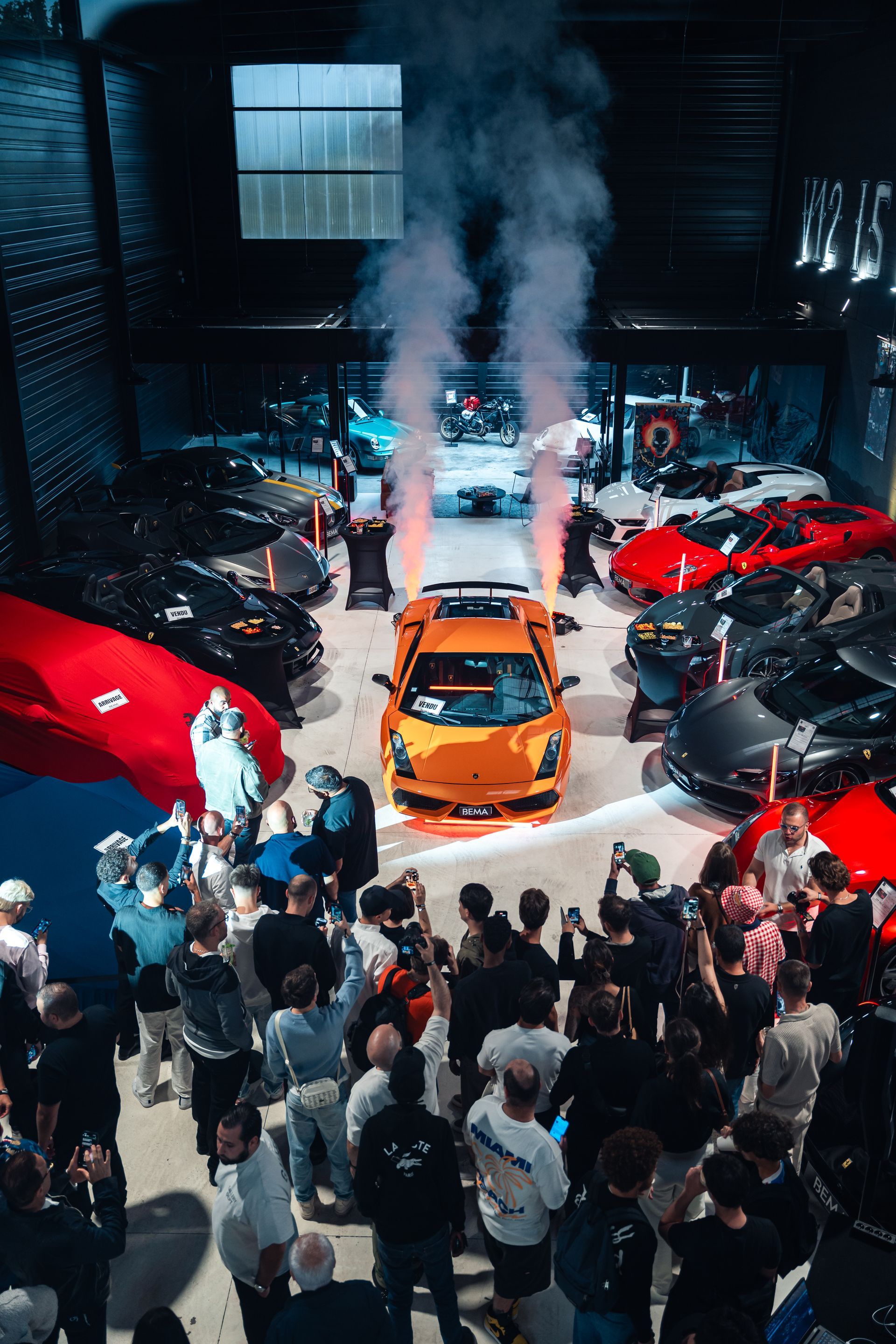 Voiture de sport orange entourée d'une foule et de fumée à l'intérieur d'un entrepôt automobile.
