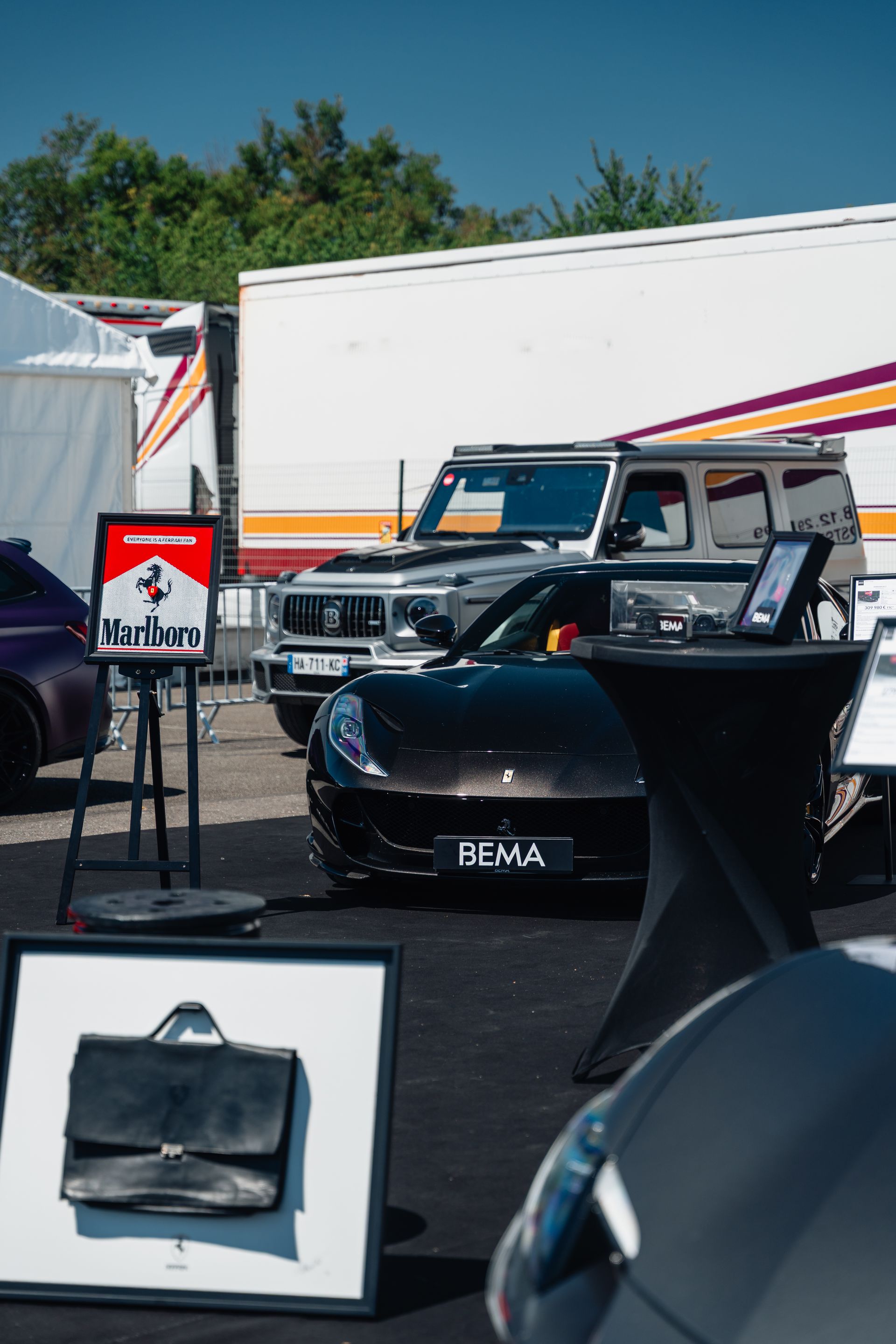 Voiture de sport noire, SUV argenté et remorque blanche lors d'un événement en plein air. Une mallette est exposée devant.