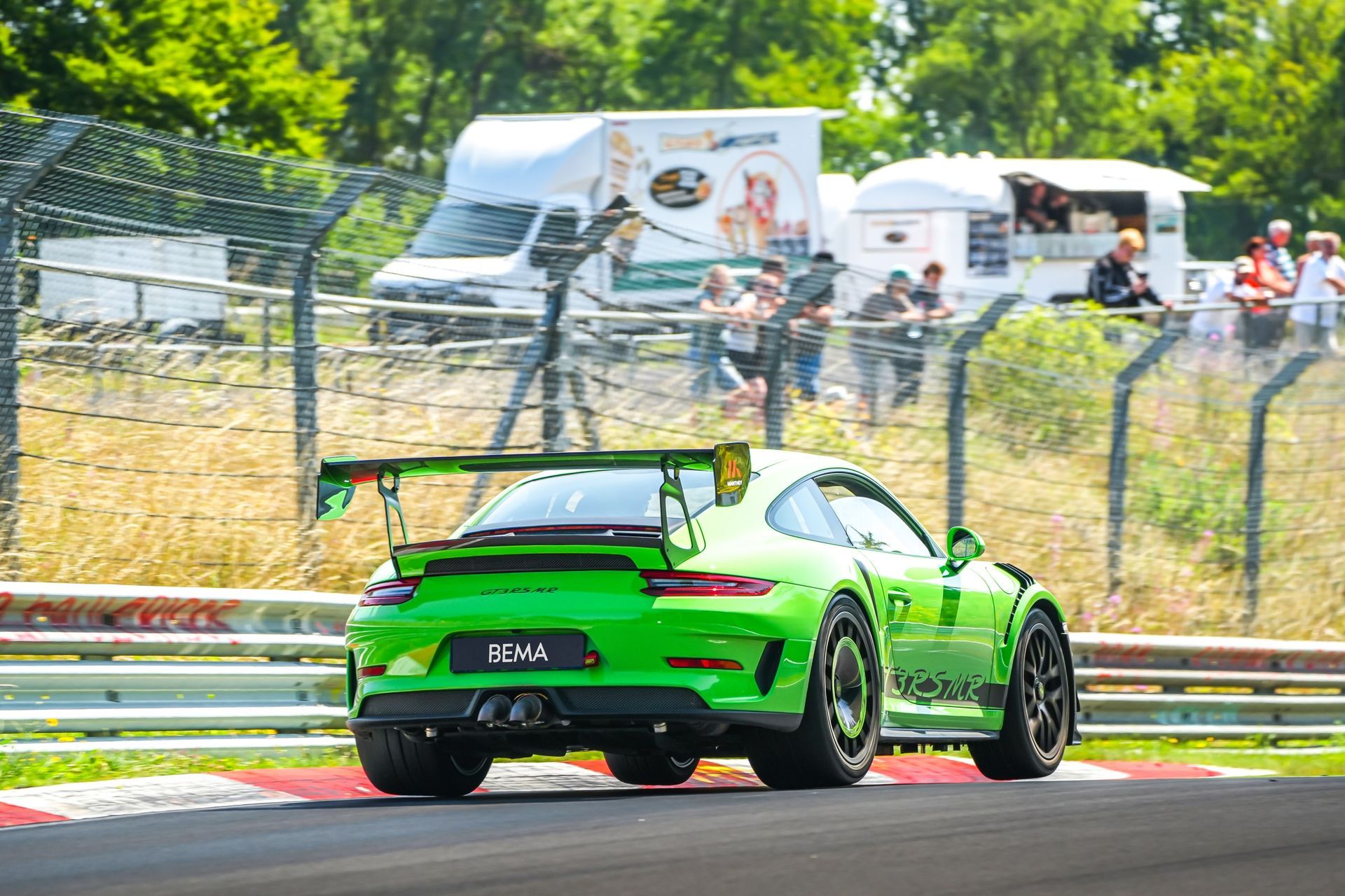 Voiture de course Porsche verte sur une piste, grand aileron, roues noires, spectateurs, food trucks en arrière-plan.