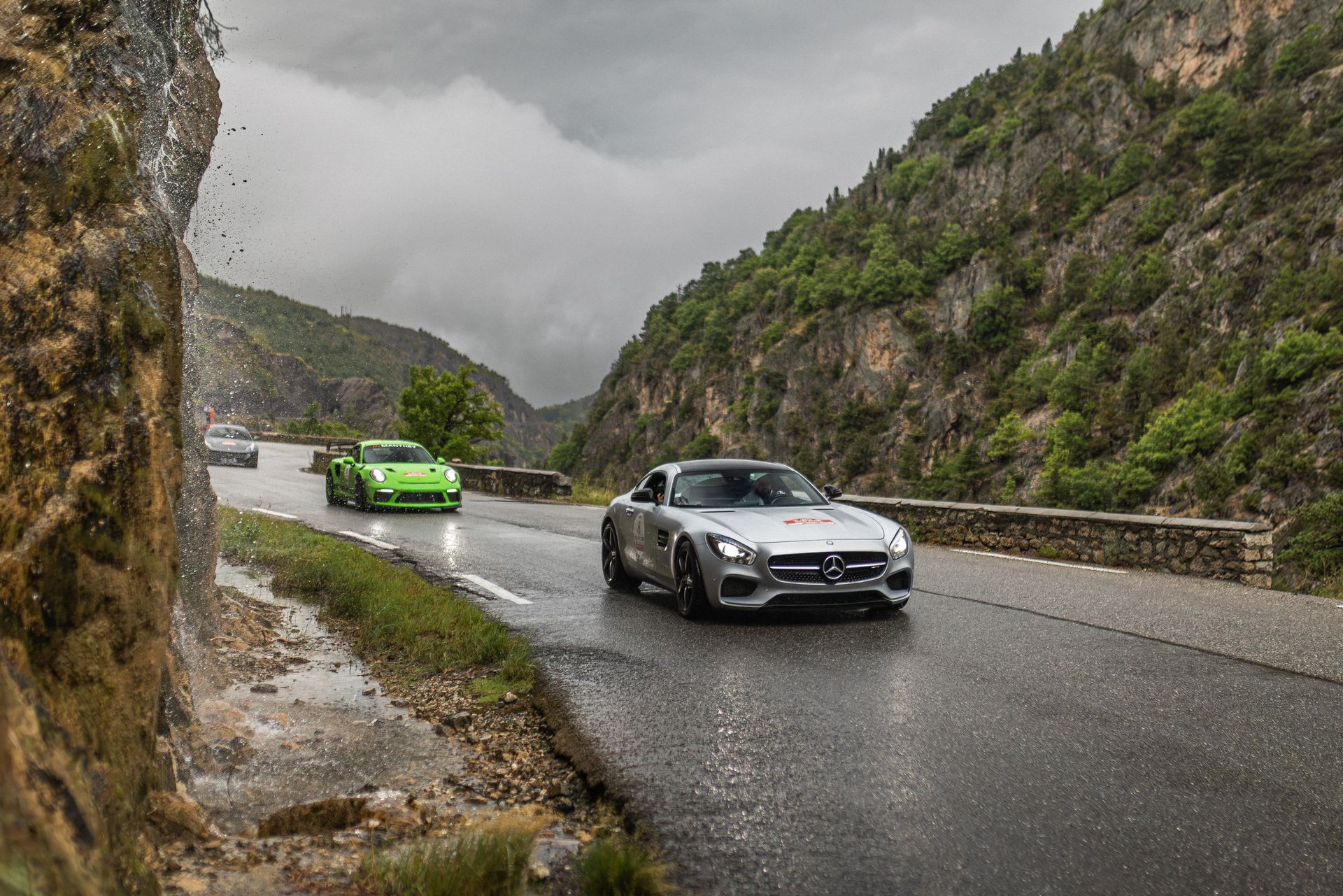 Deux voitures de sport roulent sur une route de montagne mouillée ; la voiture verte à l'arrière, la voiture argentée à l'avant.
