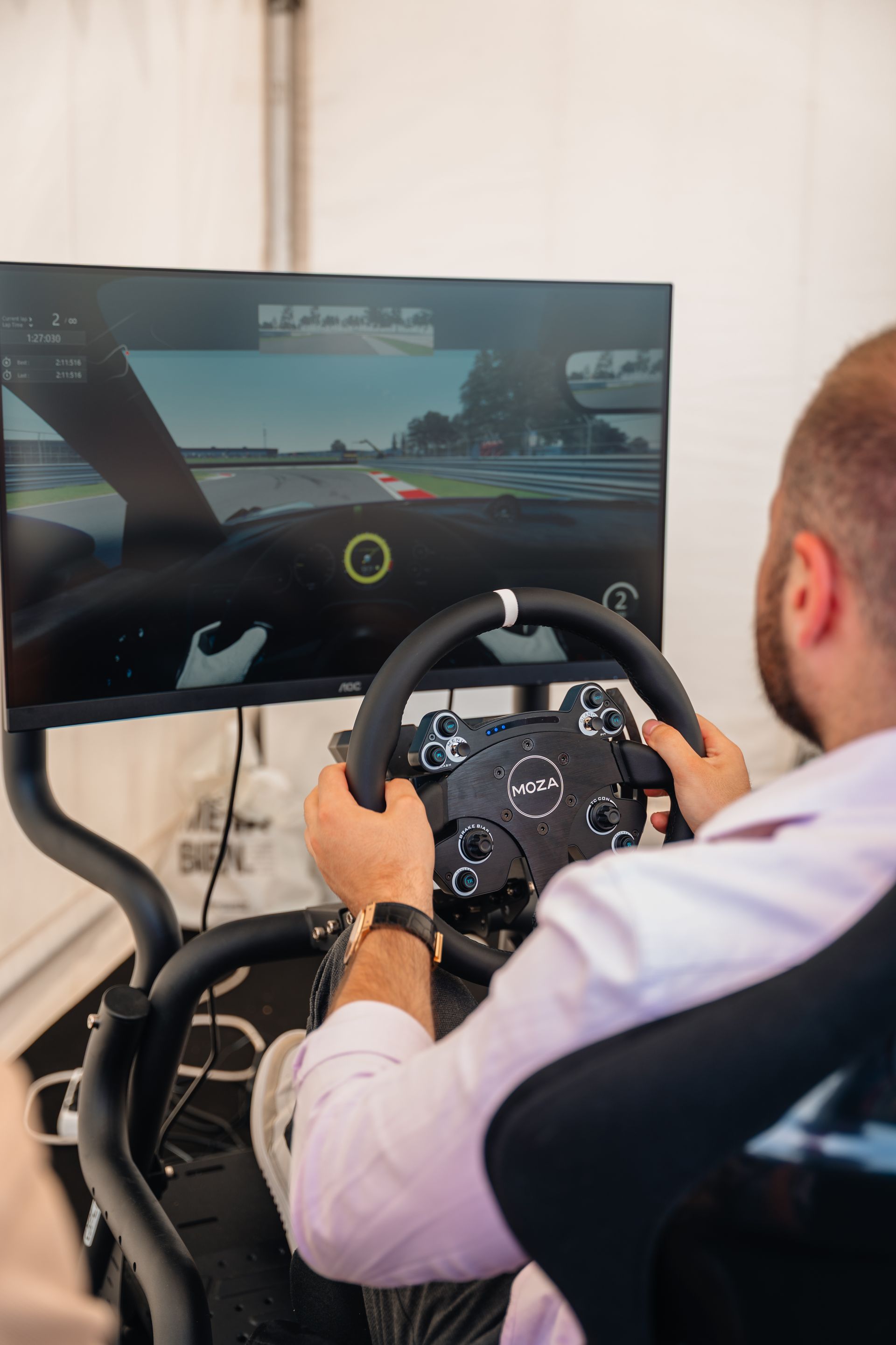 Un homme joue à un jeu vidéo de course avec un volant et un écran. Il porte un t-shirt rose clair.