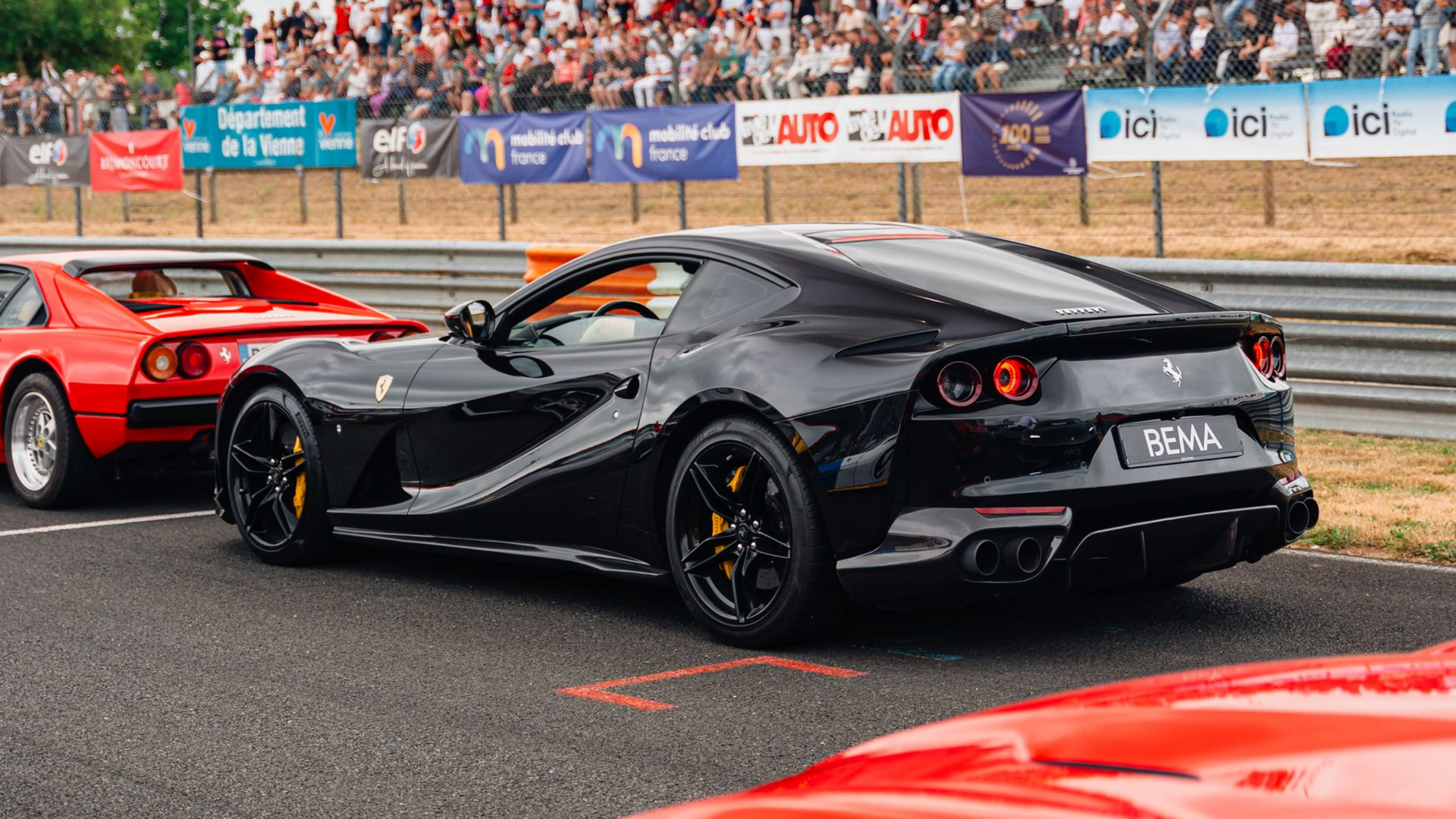 Voiture de sport Ferrari noire sur une piste de course, entourée d'autres Ferrari rouges et de spectateurs en arrière-plan.