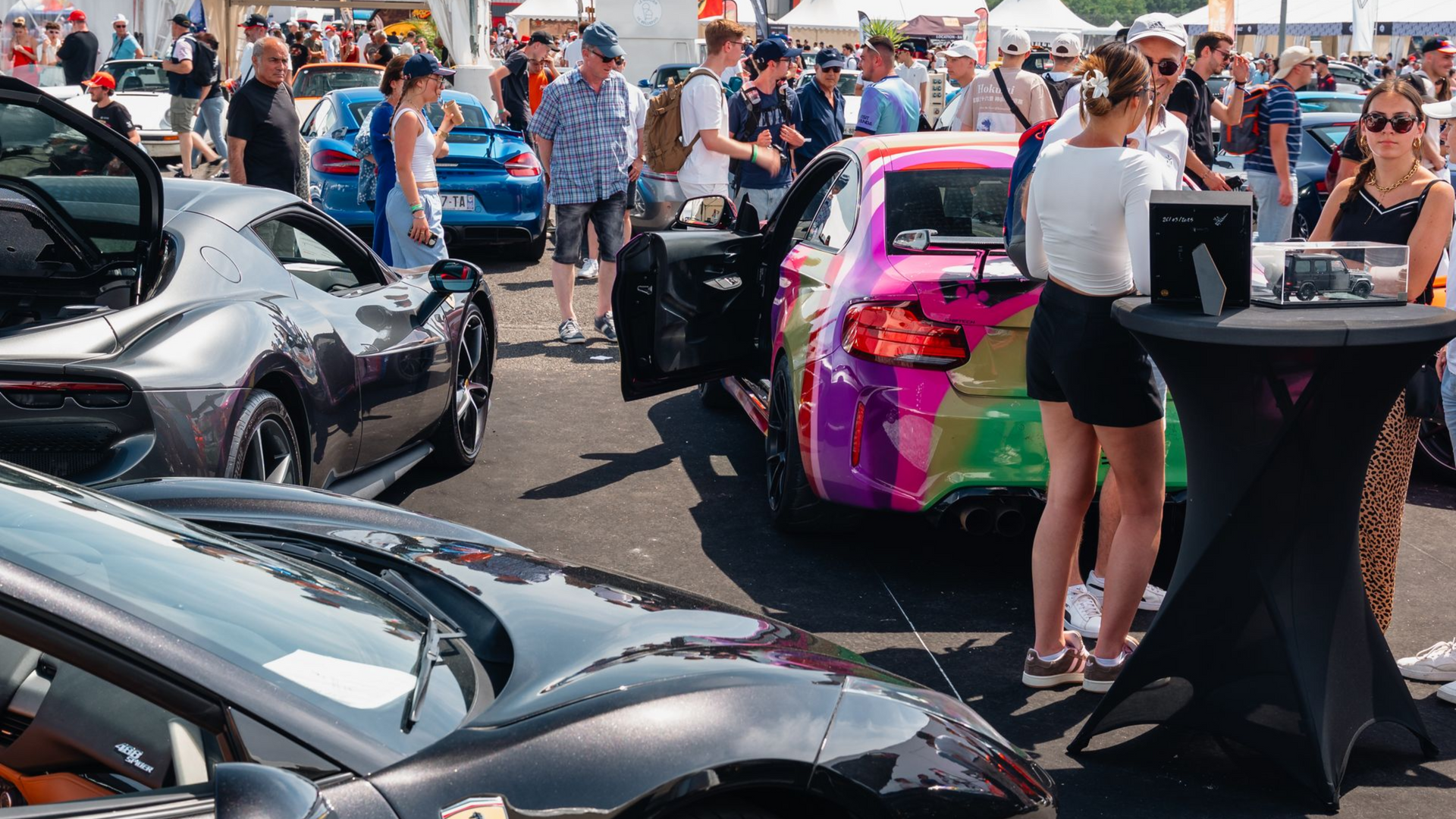Ferrari noire garée lors d'un salon automobile, avec une foule de personnes et d'autres véhicules en arrière-plan.
