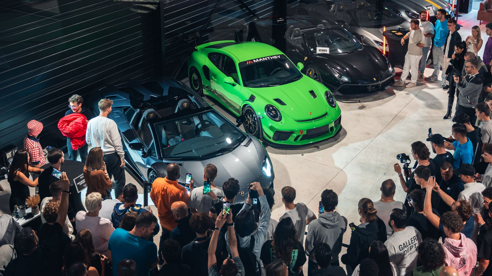 Les gens se rassemblent autour de trois voitures de sport dans une salle d'exposition ; une voiture verte vif est au centre de l'attention.