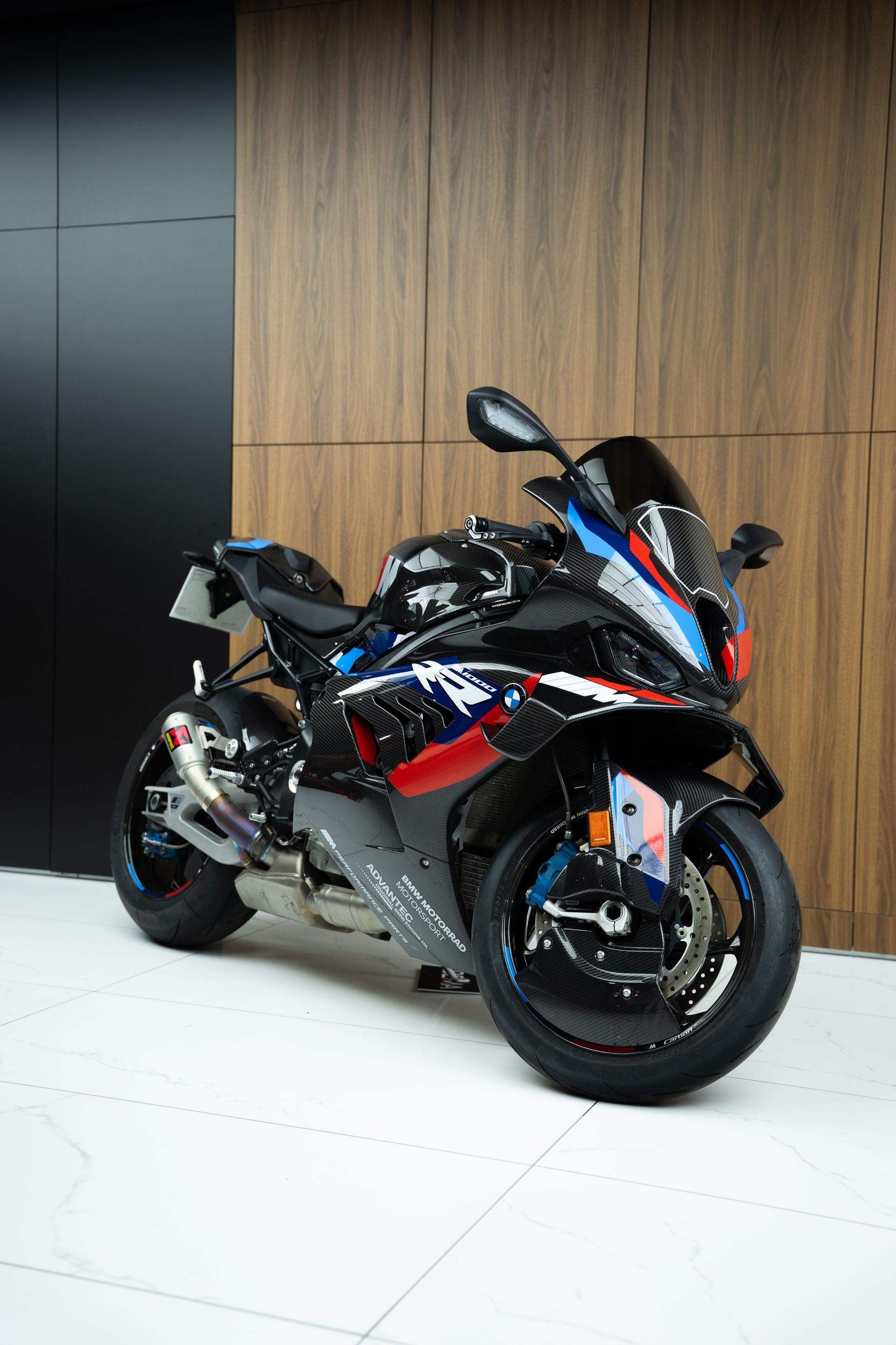 Une moto BMW noire, bleue et rouge est garée à l'intérieur, contre un mur lambrissé.