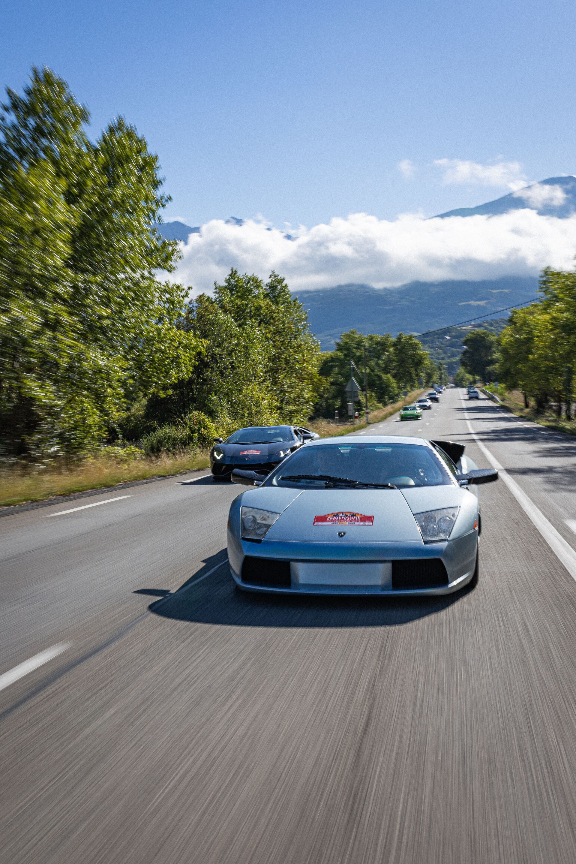 Lamborghini argentée sur une route, suivie d'autres voitures, arbres sur les côtés, montagnes en arrière-plan.