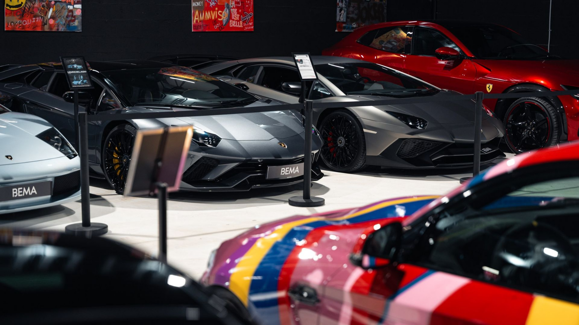 Voitures de luxe dans une salle d'exposition, dont une voiture à rayures arc-en-ciel, une Lamborghini noire et une Ferrari rouge.