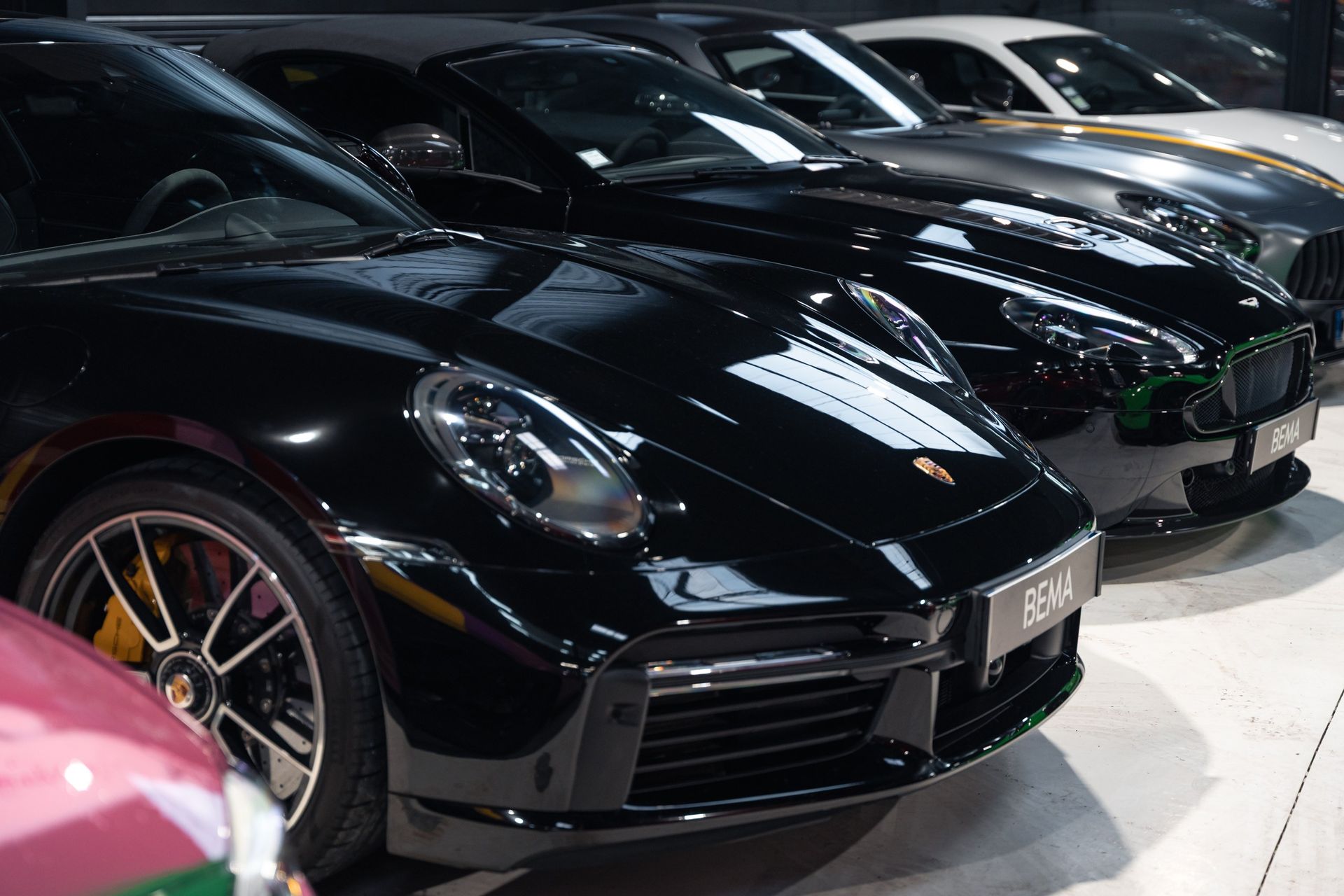 Rangée de voitures de luxe, dont une Porsche noire, garée à l'intérieur d'une salle d'exposition.